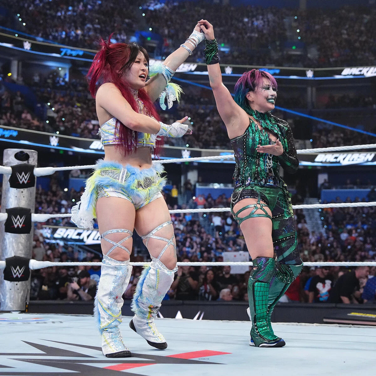 Iyo sky and asuka - the game Two female WWE wrestlers celebrate a victory in the ring, raising their arms together as the crowd cheers in the background.