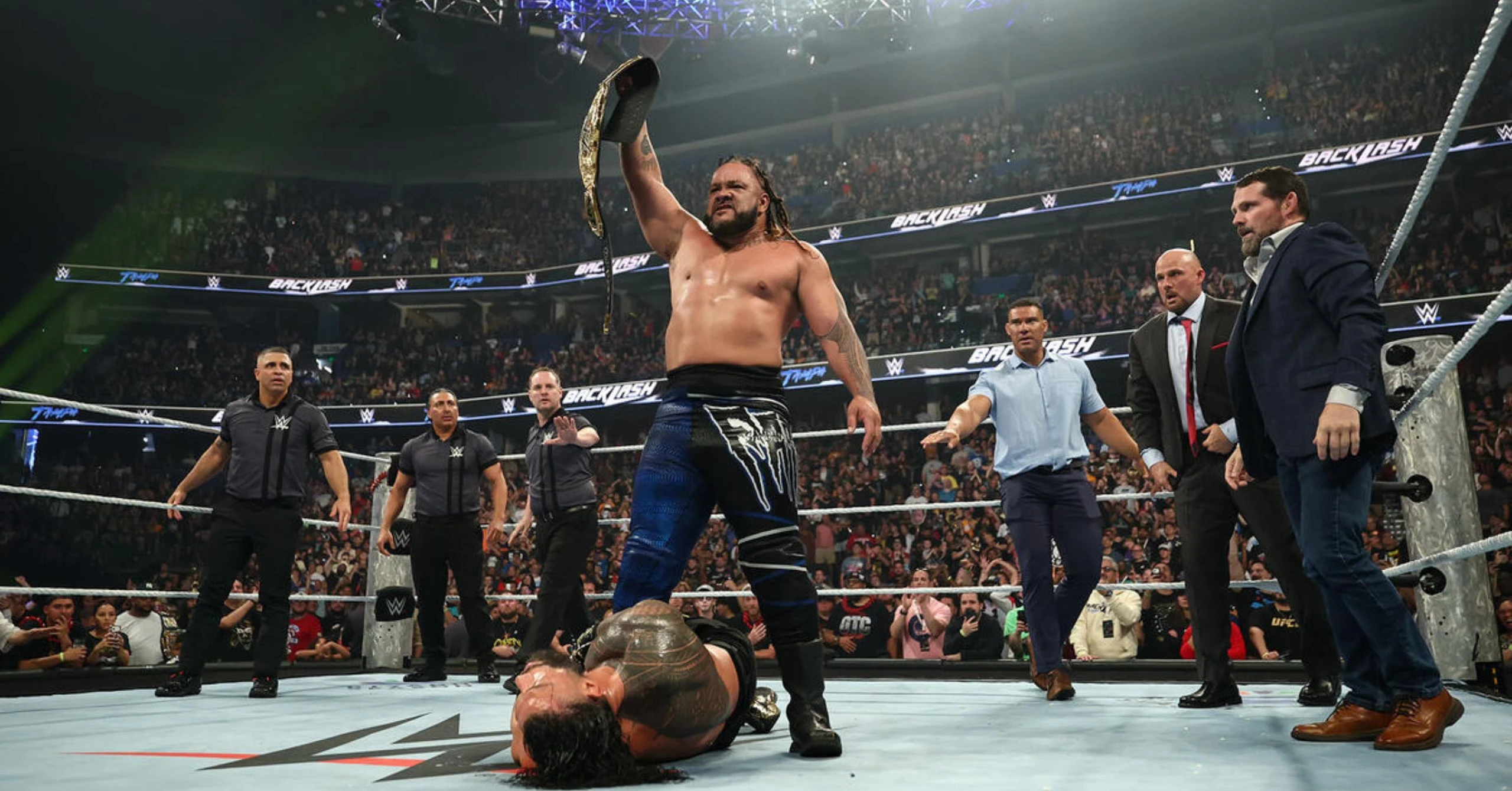 WWE wrestler raises championship belt in the ring as the crowd roars, an opponent lies on the mat nearby.