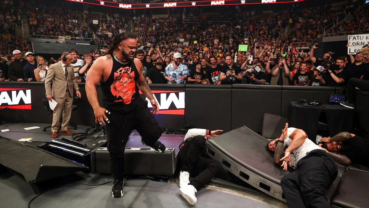 WWE-style arena moment: a muscular wrestler in a black sleeveless shirt walks past a toppled barrier toward a fallen opponent as a packed crowd watches from the stands.