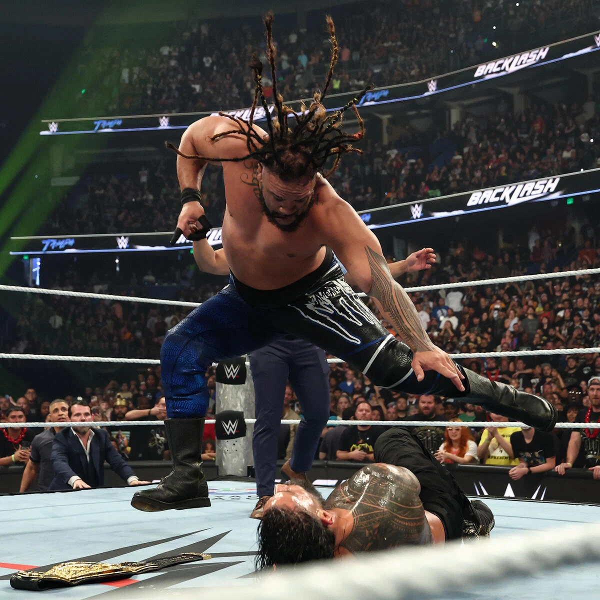 Jacob fatu vs roman - the game WWE wrestler with long dreadlocks leaps over a downed opponent inside the ring as the crowd watches, with a championship belt on the mat nearby.