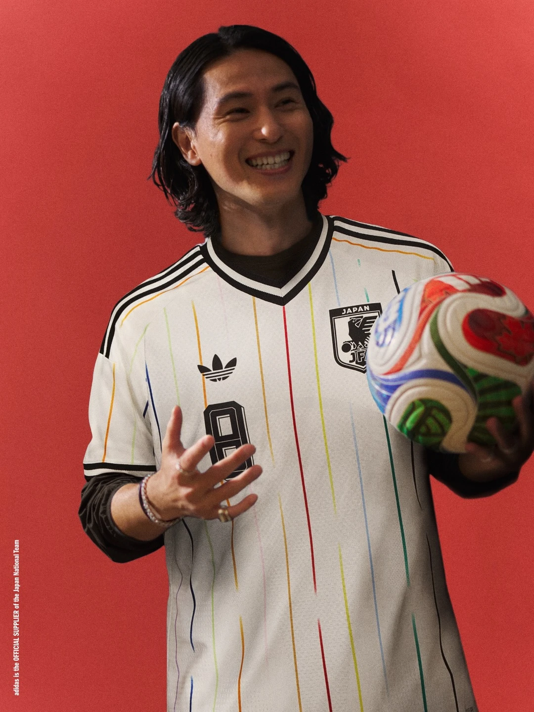 Japan away - the game Smiling person in a white Japan national team jersey with rainbow stripes, holding a colorful soccer ball against a red background.