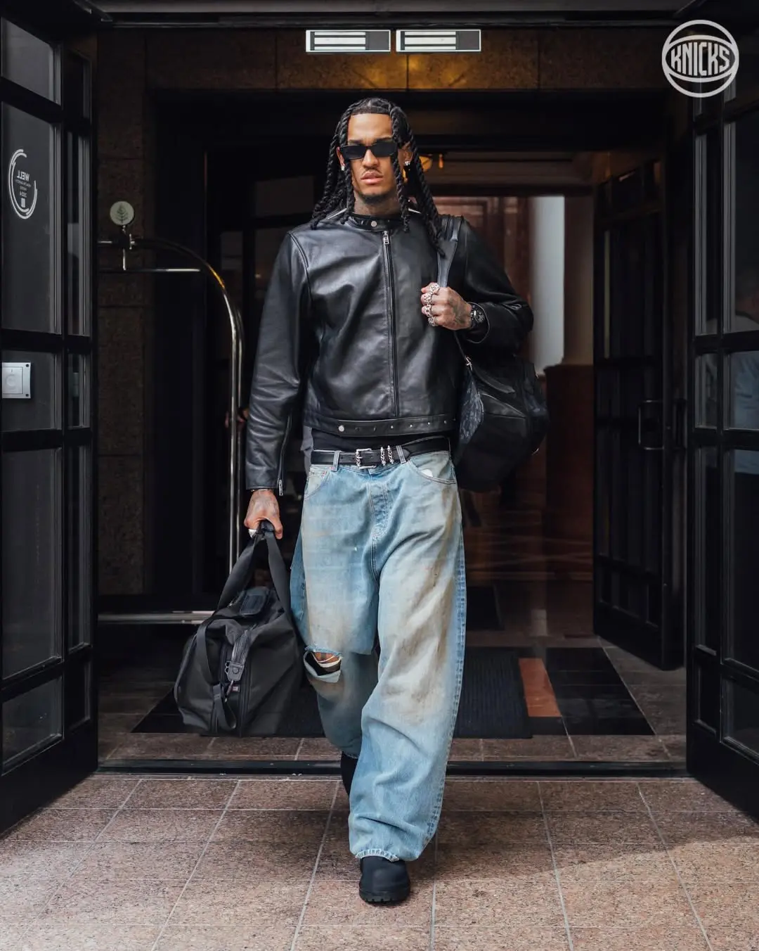 Jordan clarkson - the game Stylish man in a black leather jacket with a bag slung over his shoulder, wearing oversized distressed jeans and sunglasses as he exits a building.