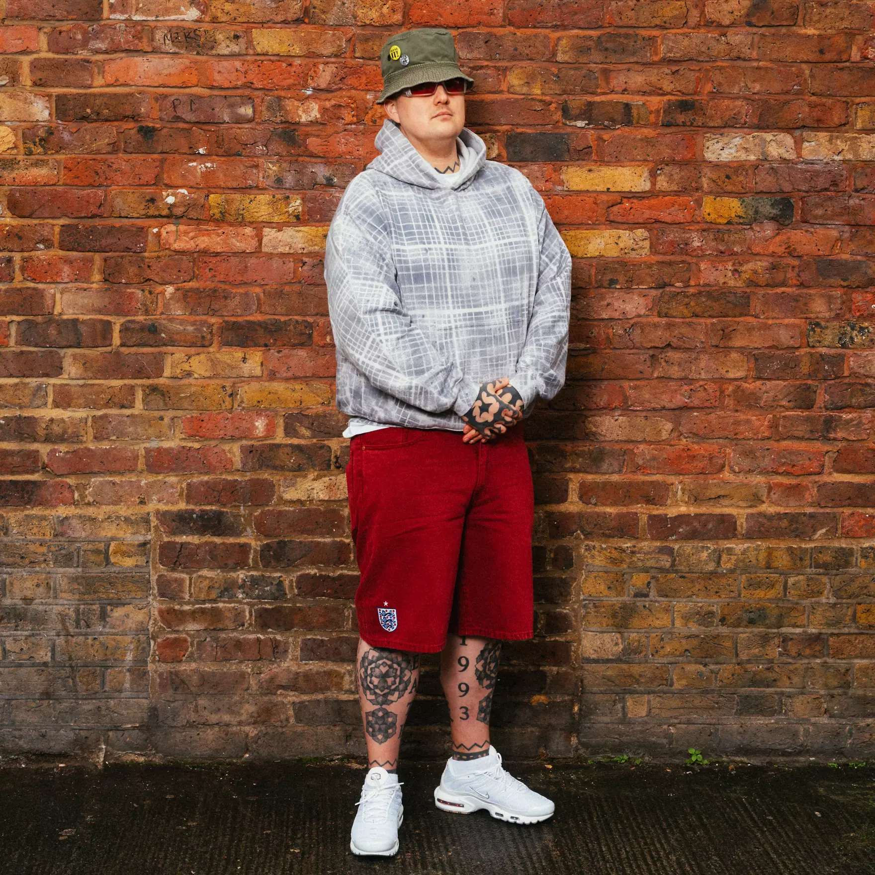 Jorts - the game Person standing against a brick wall wearing a gray plaid hoodie, red shorts, white sneakers, and a green bucket hat with pins; visible tattoos on legs.