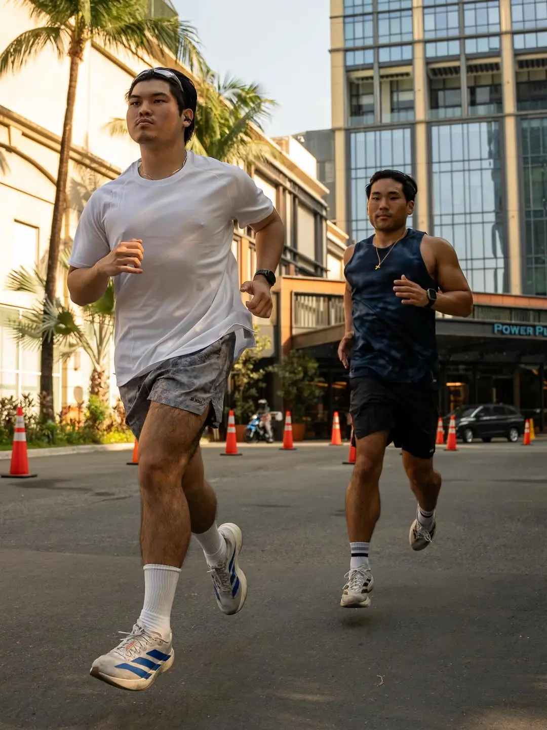 Two men are running on a city street with orange traffic cones and tall glass buildings in the background.
