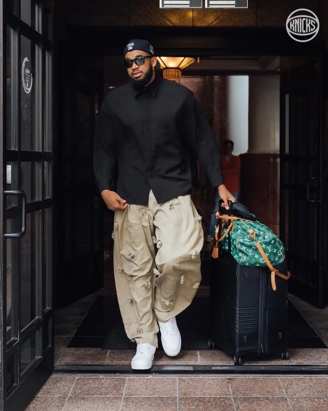 Kat - the game Man in a black button-up shirt, beige distressed cargo pants, white sneakers, cap and sunglasses exits a doorway pulling a black rolling suitcase with a green Louis Vuitton duffel on top.