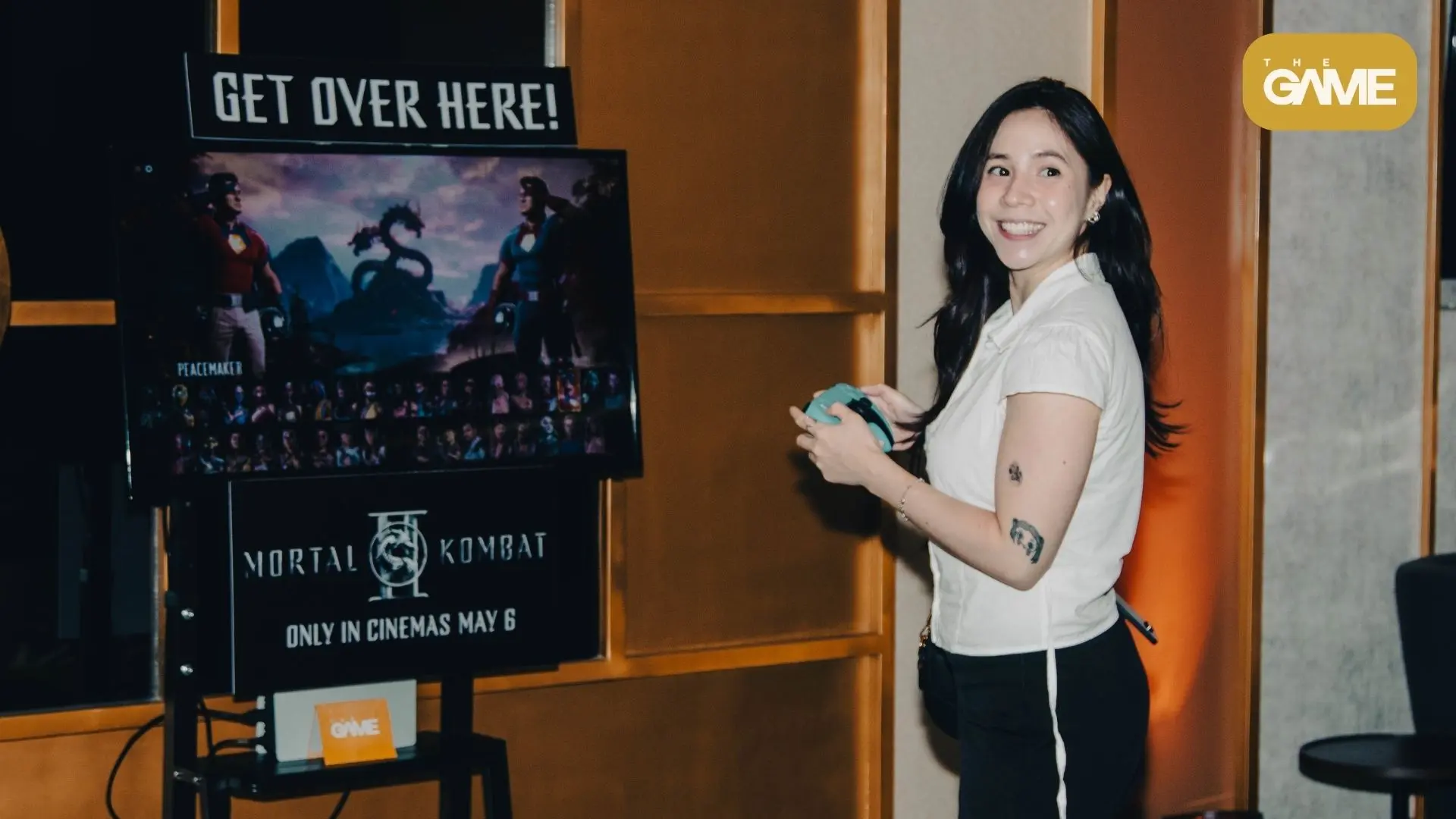 Kafi - the game Smiling woman holds a teal game controller beside a Mortal Kombat poster that reads GET OVER HERE! and 'Only in cinemas May 6' in the background, with The Game logo top-right.