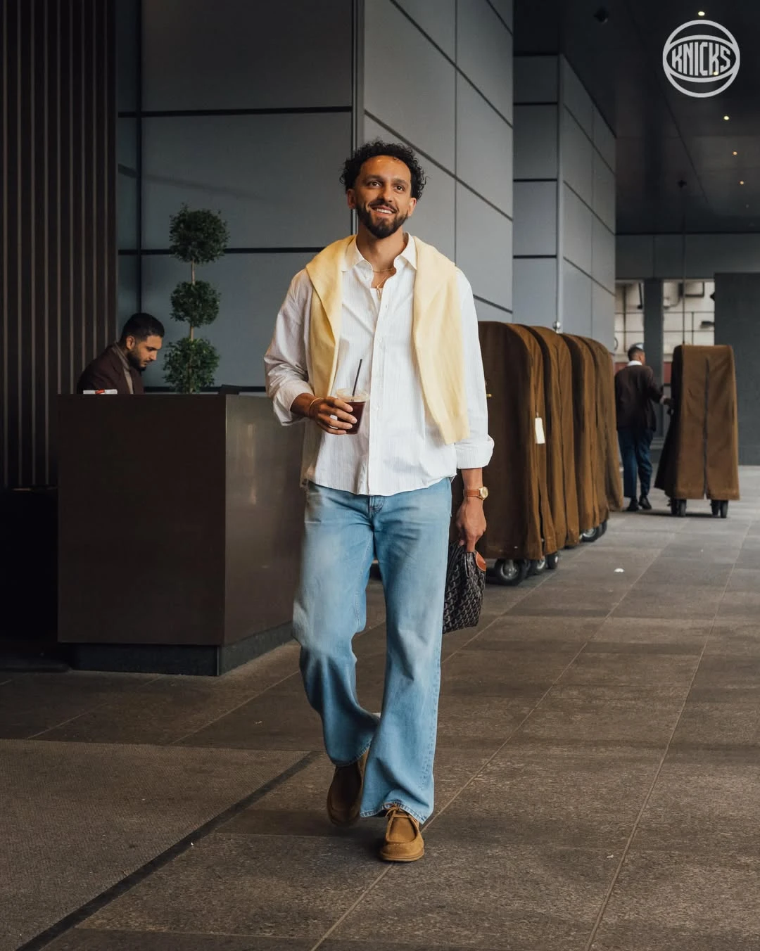 Landry shamet - the game Man walking through a modern lobby with a drink in one hand and a light sweater draped over his shoulders, carrying a patterned bag.