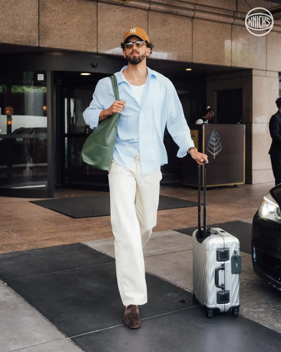 Landry shamet - the game Casual male traveler wearing a light blue shirt, white pants, brown cap and sunglasses, pulling a silver rolling suitcase outside a hotel entrance with a green tote over his shoulder.