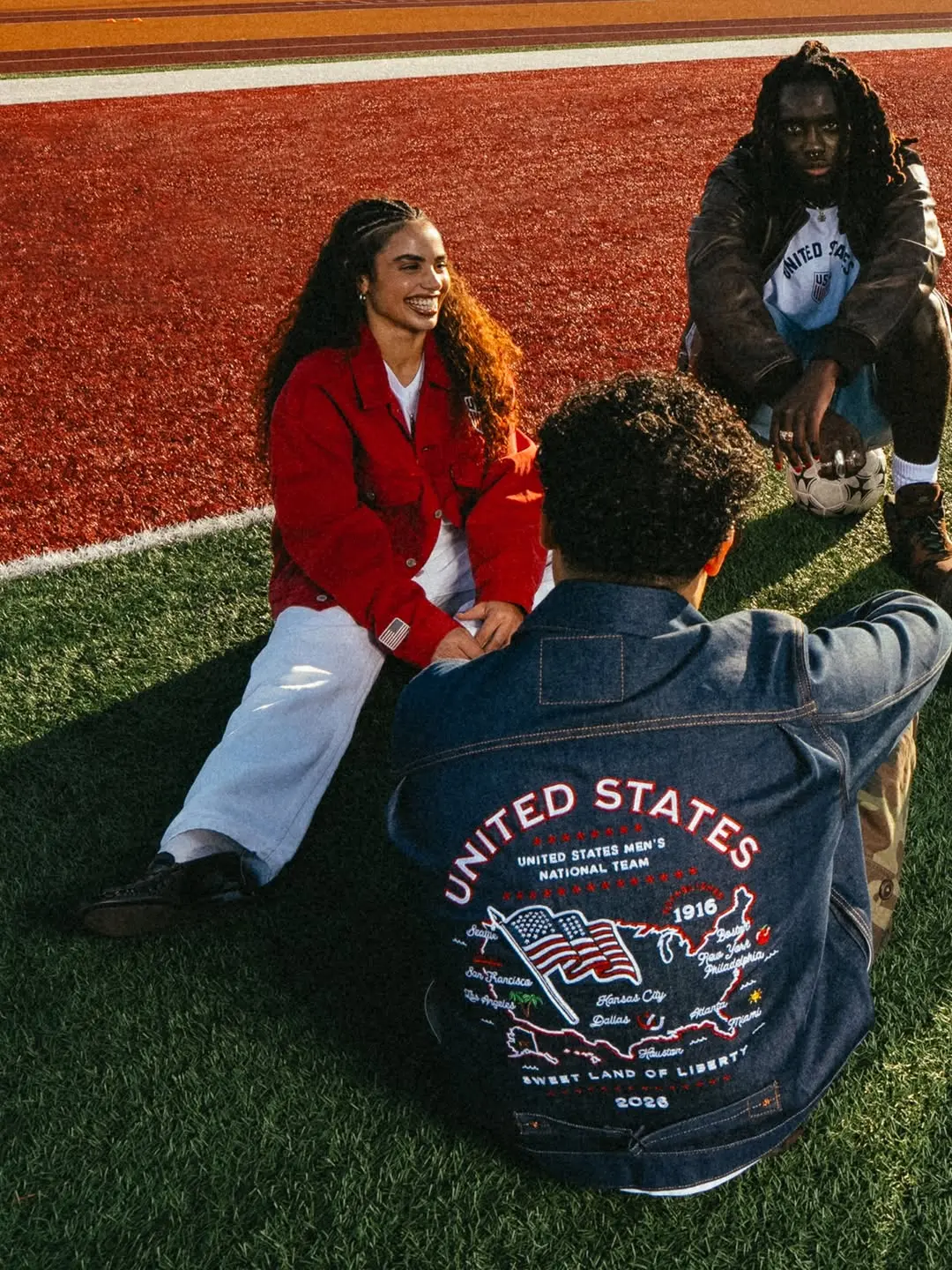 Three friends sit on artificial turf near a red track, chatting and smiling.