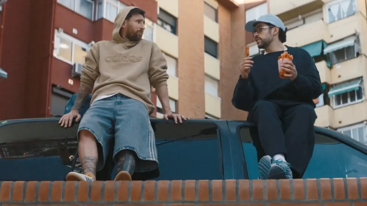 Messi and bad bunny - the game Two men sit on a car in an urban street, chatting; one wears a beige hoodie and denim shorts, the other with a cap holding snacks and a drink.