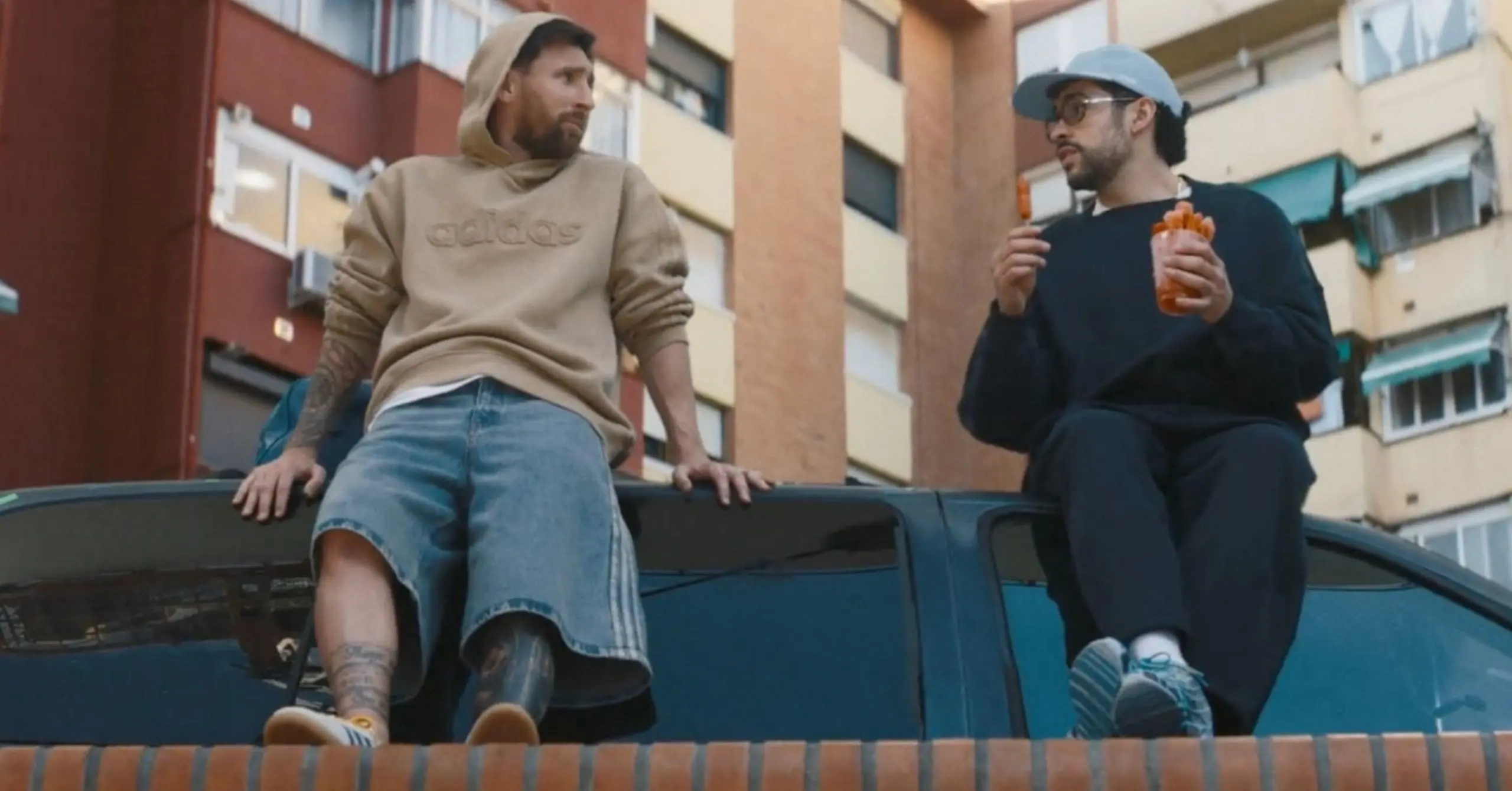 Two men sit on the hood of a dark car in a city street; left man in a beige hoodie and denim shorts, right man in a cap and glasses holding an orange drink and snacks.