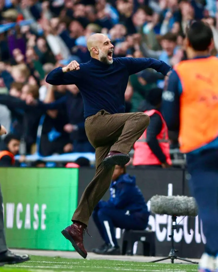 Pep guardiola - the game Bearded man in a navy shirt and brown trousers leaps mid-air with a fist pump on the sideline, stadium crowd cheering in the background.