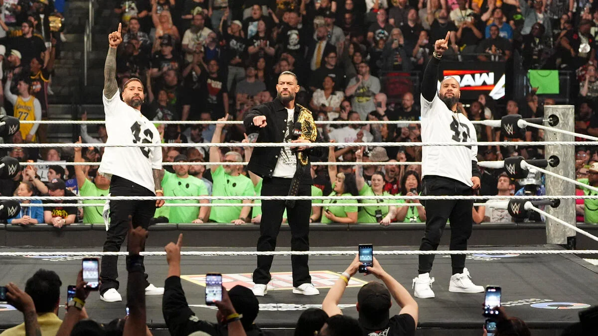 Three wrestlers stand in the ring; the center figure wears a championship belt while two others in white shirts point upward as the crowd cheers.