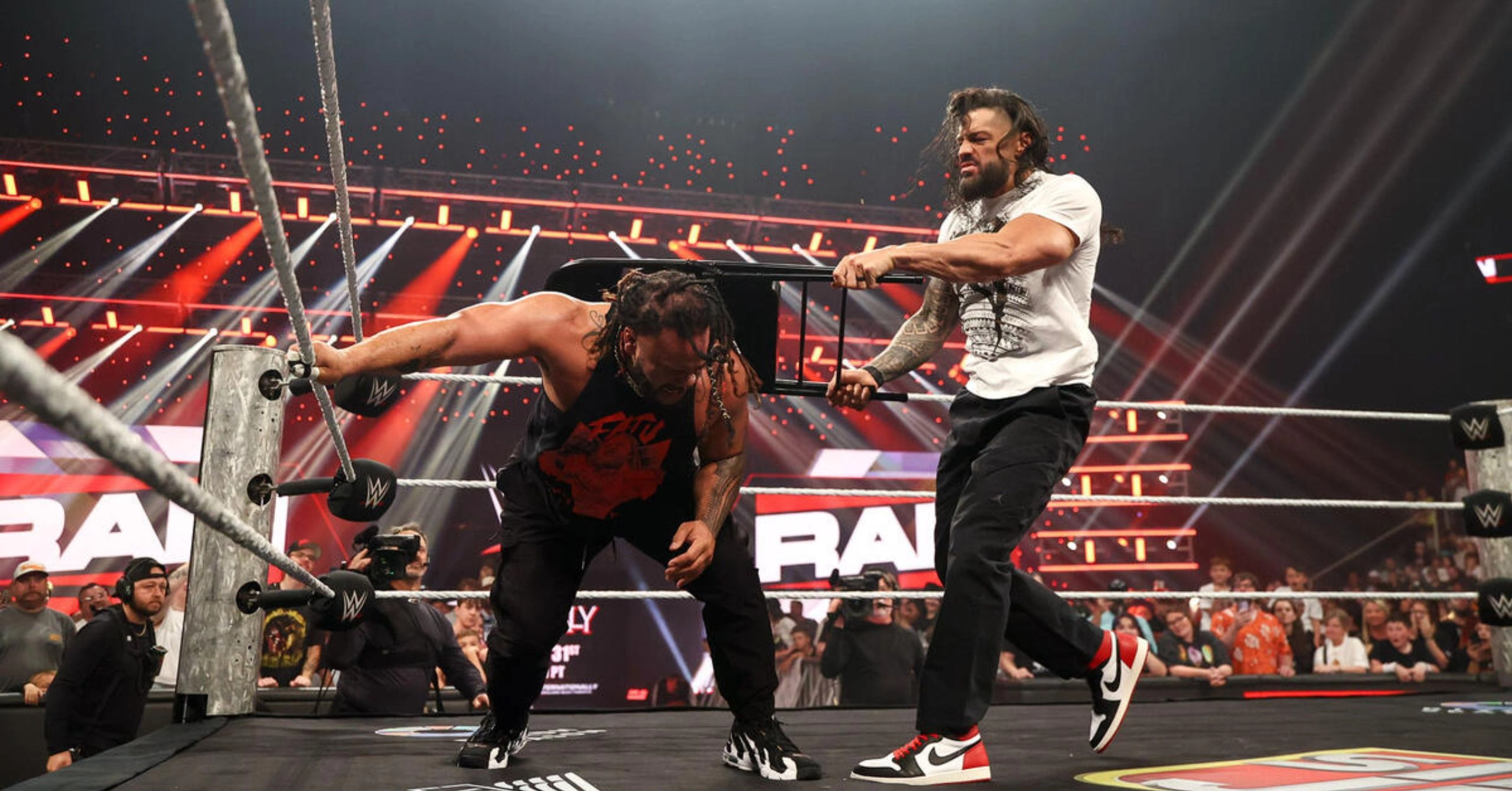 Two muscular wrestlers clash in a WWE ring during a RAW match, one gripping a chair while the other leans forward amid a cheering crowd and bright arena lights.