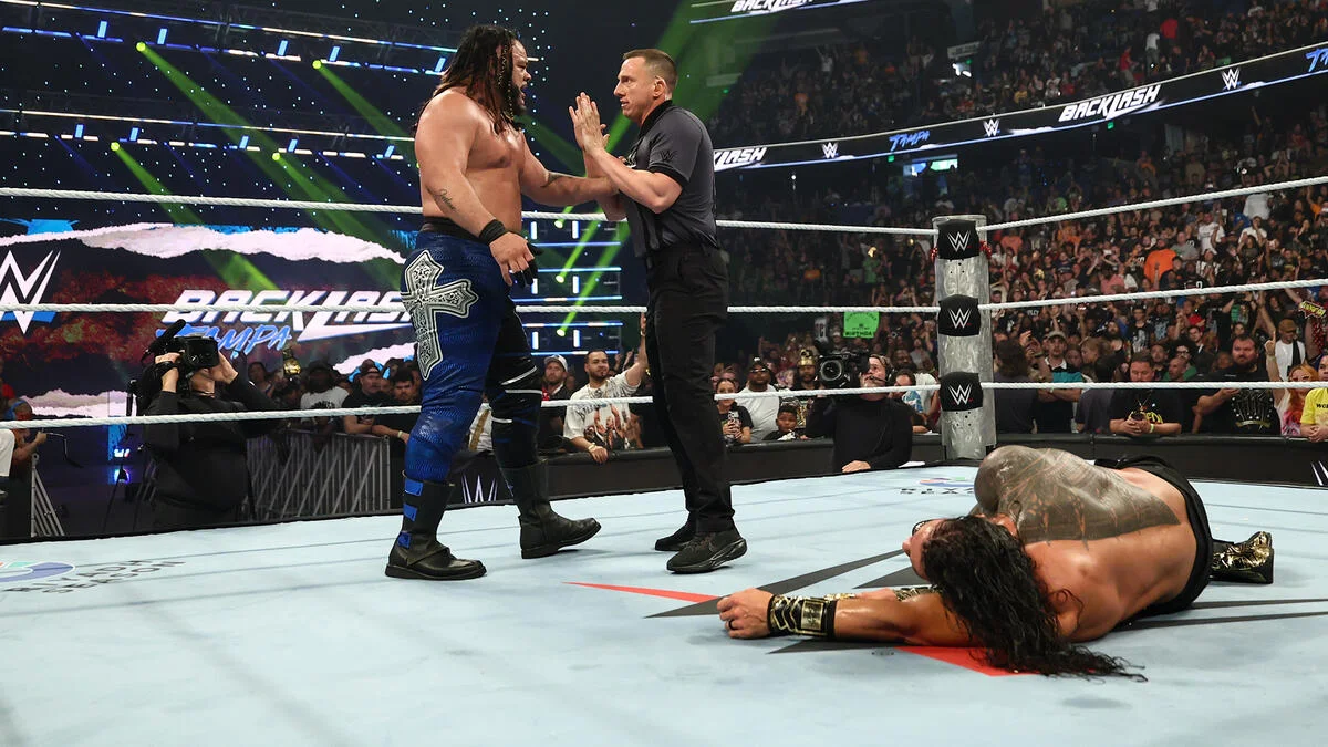 WWE ring moment: two wrestlers stand facing each other as a referee gestures, while a third wrestler lies on the mat nearby and a crowd watches.