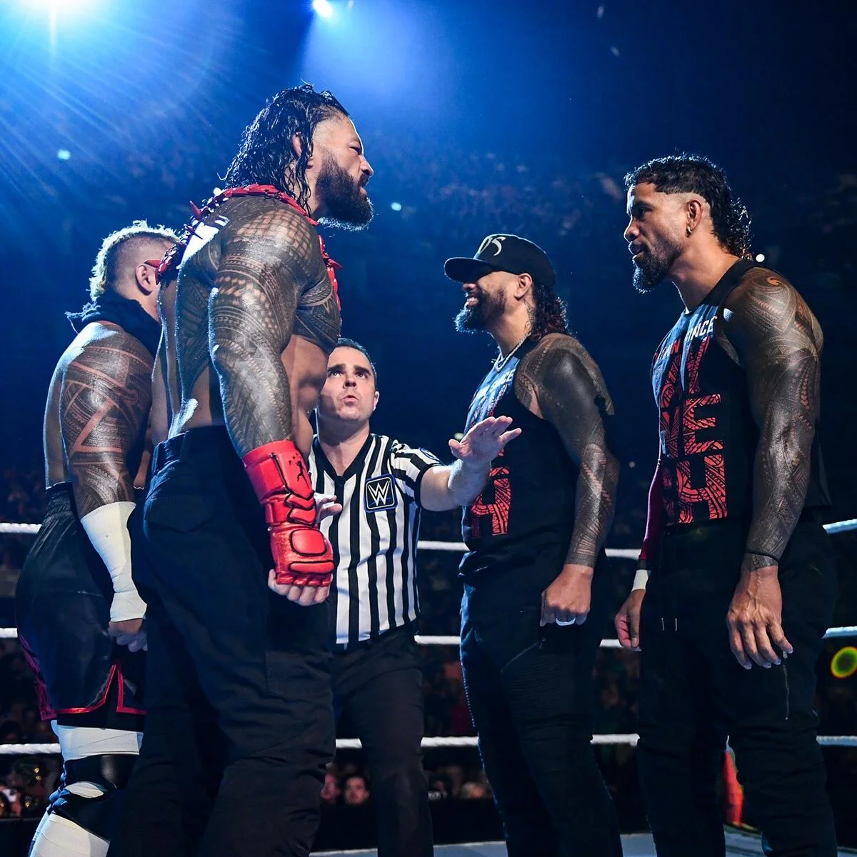 Four professional wrestlers stand nose-to-nose in a wrestling ring, with a striped-shirt referee between them under bright arena lights.
