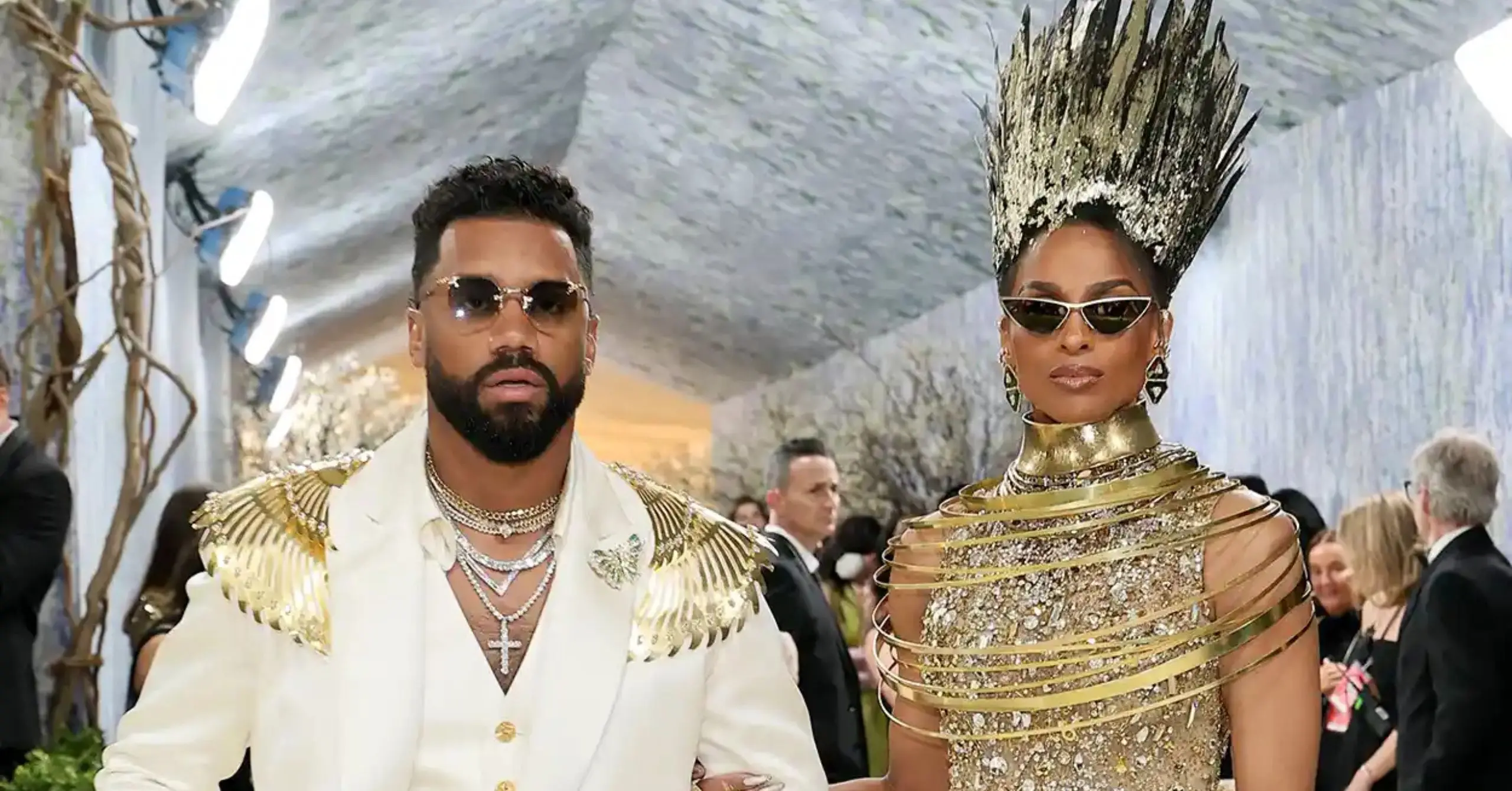 Two fashion-forward individuals walk a glamorous red carpet; man in white suit with gold accents and jewelry, woman in a metallic gold ensemble with spiky hair and sunglasses.