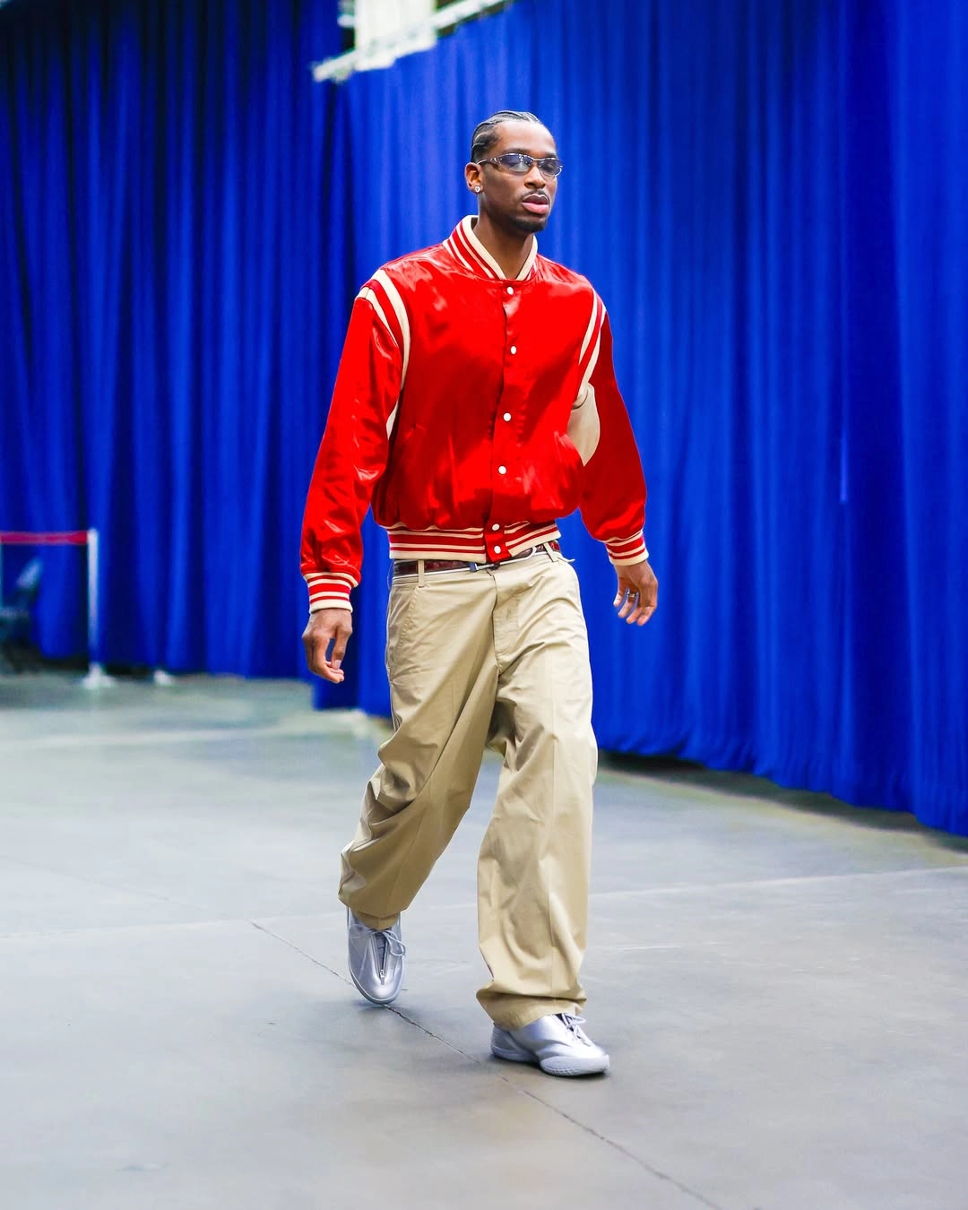 Sga - the game Professional basketball player Shai Gilgeous-Alexander strides through a sports venue tunnel showcasing a vibrant retro look. He pairs a glossy, satin red varsity bomber jacket featuring cream-colored striped trim with oversized, wide-leg beige khaki trousers. His look is styled with silver metallic low-top sneakers and sleek, narrow-rimmed sunglasses.