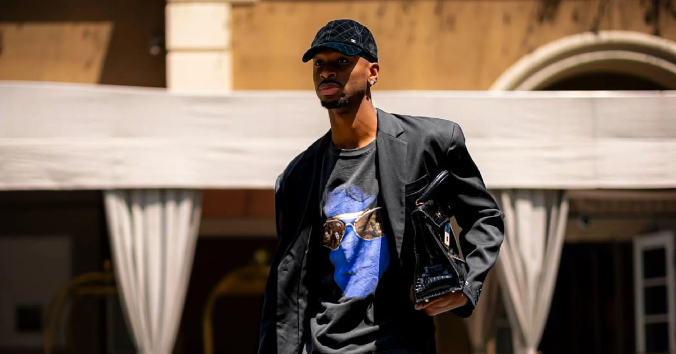 Man wearing a black cap, black jacket, and graphic tee walking outdoors in an urban setting with a beige building behind him.