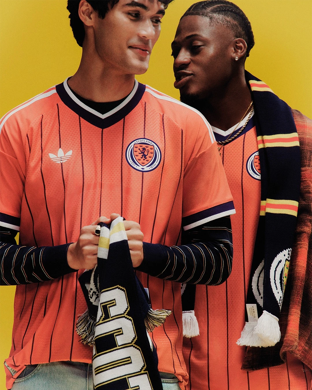 Scotland away - the game Two people in orange Scotland football jerseys stand against a bright yellow background, one adjusting a scarf and both looking toward each other.
