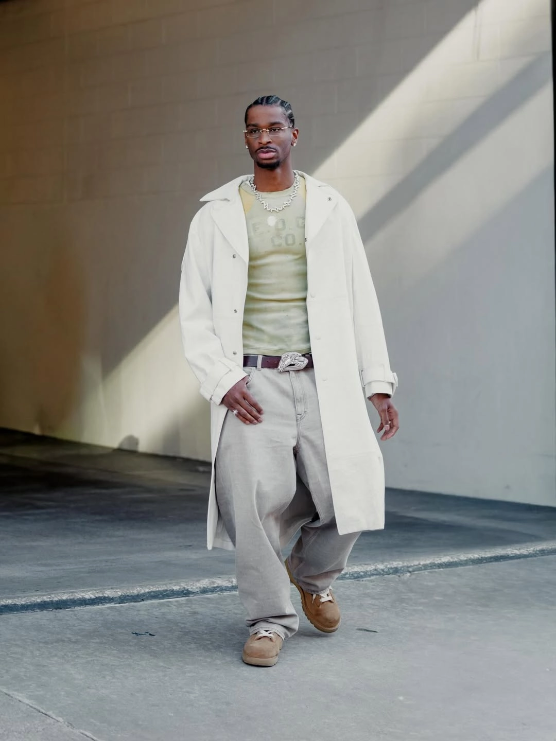Shai - the game Description: Professional basketball player Shai Gilgeous-Alexander showcases a curated neutral outfit while arriving at an arena tunnel. He wears an oversized ecru trench coat layered over a fitted, olive-green graphics t-shirt, paired with ultra-baggy light grey denim jeans. Accessories include clear-framed glasses, a custom iced-out signature logo chain, a large statement belt buckle, and brown suede shoes.