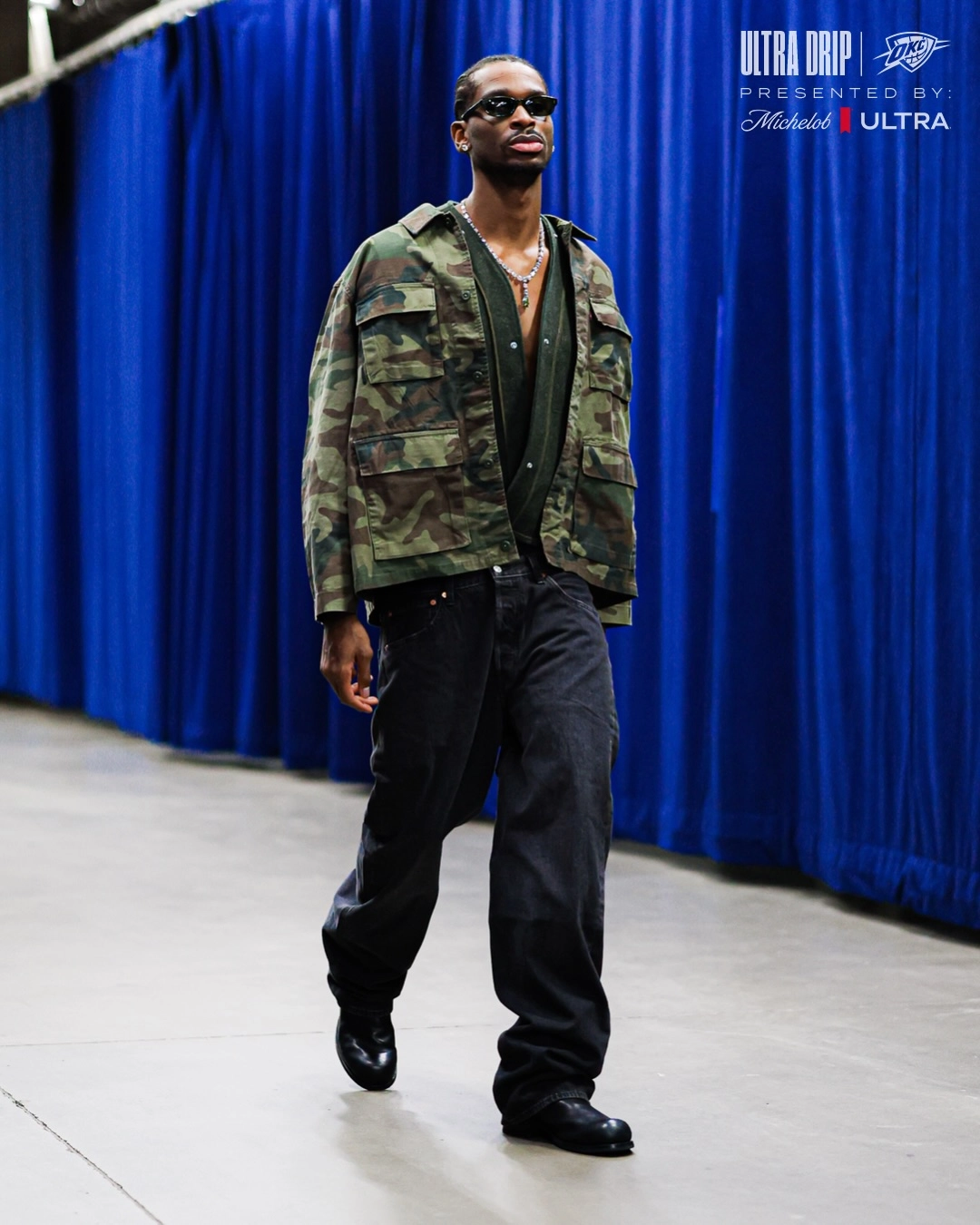 Shai gilgeous-alexander - the game Professional basketball player Shai Gilgeous-Alexander showcases a rugged luxury look while walking through an arena tunnel. He wears an oversized woodland camouflage field jacket over a low-cut dark olive green knit shirt. The top is paired with loose, baggy washed black denim jeans and heavy-duty black leather boots. He accessorizes with dark rectangular sunglasses and a statement silver link necklace.