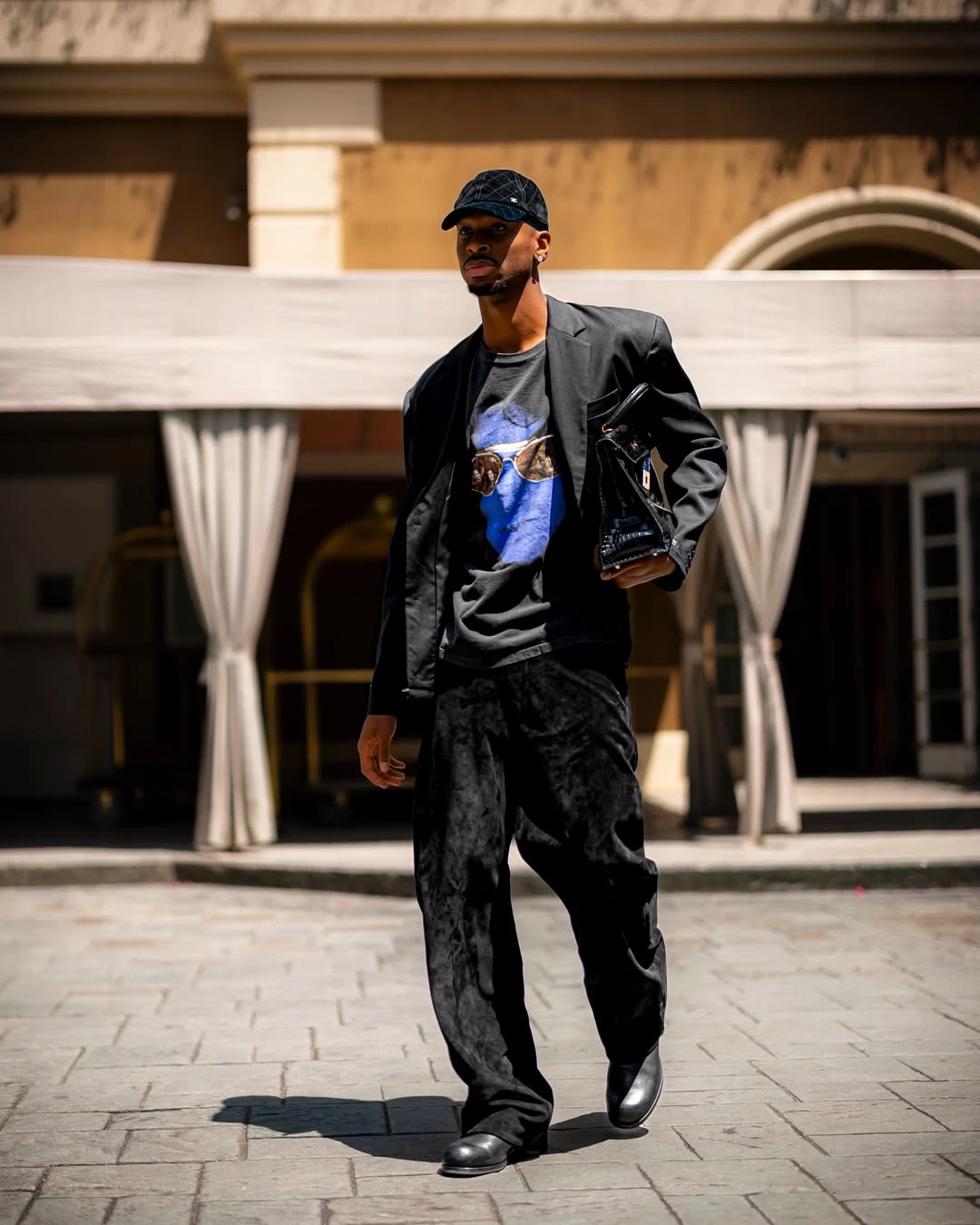 Shai gilgeous-alexander - the game Man in a black blazer and velvet pants walks on a paved courtyard, wearing a cap and carrying a black bag.