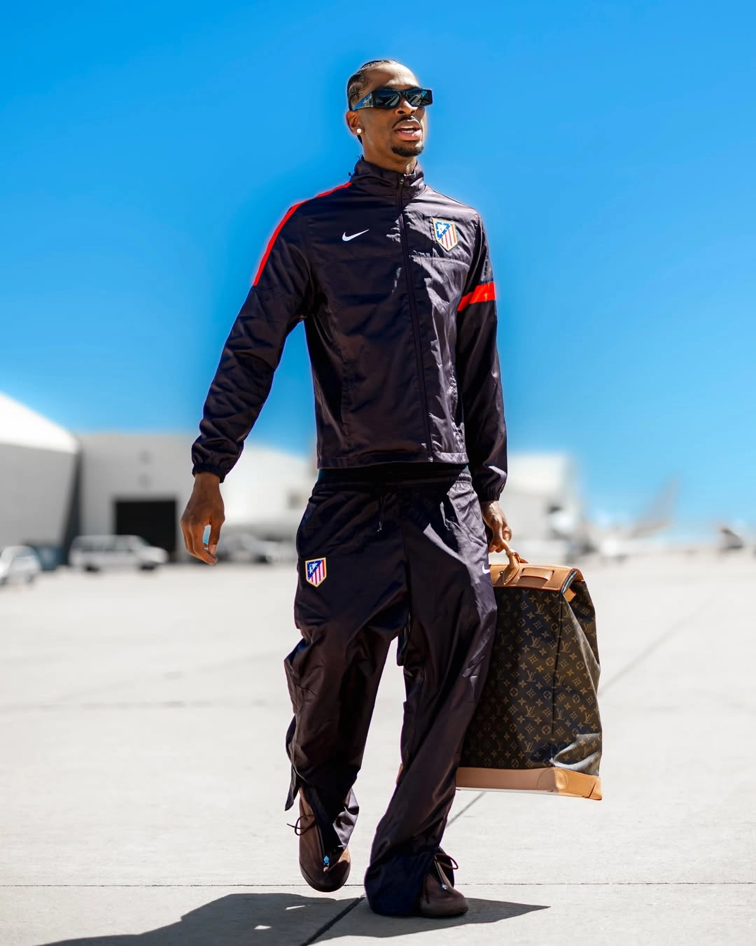 Shai tracksuit - the game Professional basketball player Shai Gilgeous-Alexander walks across an airfield runway wearing an athletic blockcore outfit. He pairs a sleek, oversized black Nike Atlético de Madrid warmup track jacket with matching loose-fitting woven tracksuit pants. He blocks out the sun with thick black rectangular sunglasses and completes his luxury travel look with a vintage Louis Vuitton Monogram canvas Steamer 45 travel bag.
