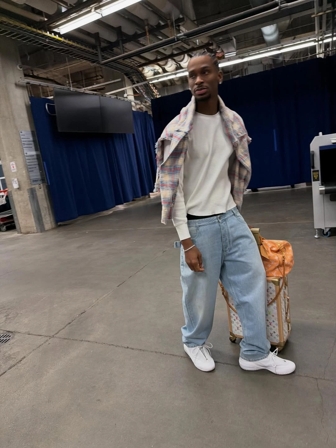 Shai w plaids - the game Professional basketball player Shai Gilgeous-Alexander arrives at an arena venue holding high-end travel gear. His comfortable, multi-layered look features a lightweight off-white thermal long-sleeve shirt paired with an intentionally draped, multi-color pastel plaid flannel over his shoulders. He coordinates the top with ultra-loose, light-wash baggy denim jeans and clean white low-top sneakers. He stands alongside his luxury travel essentials, including a Louis Vuitton rolling suitcase trunk and an orange Louis Vuitton monogram duffle bag.
