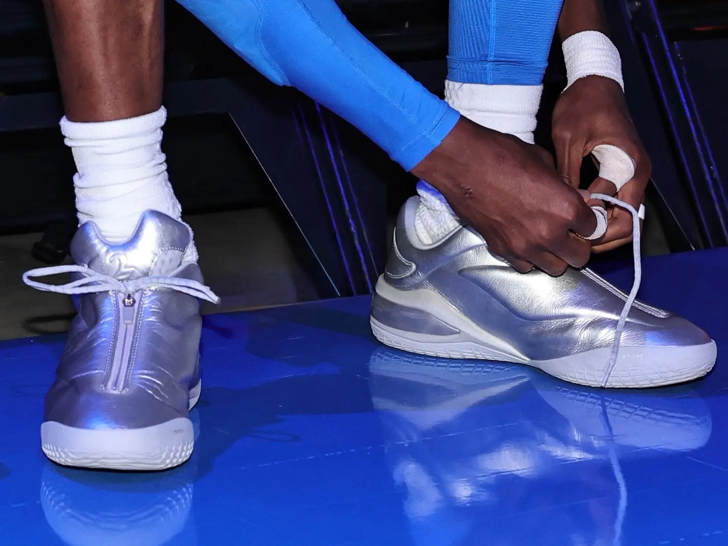 Shai - the game Person tying the laces of metallic silver athletic shoes while seated, wearing white socks on a blue floor.