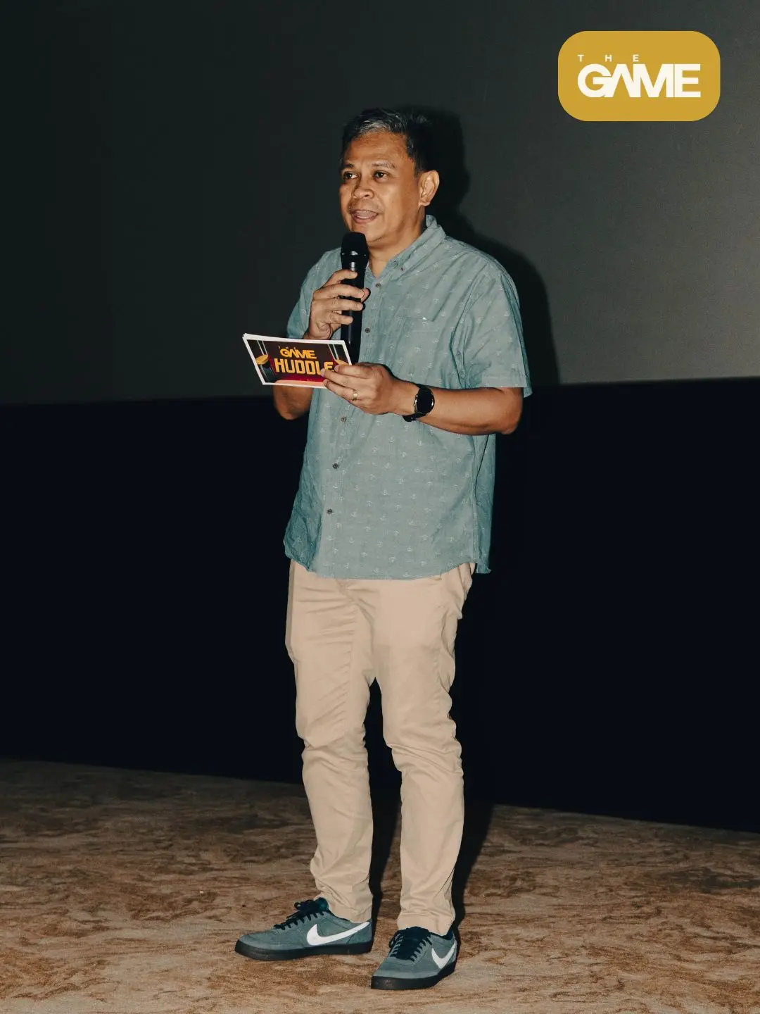 Sid - the game Man on stage speaking into a microphone while holding a cue card that says 'GAME HUDDLE', with 'THE GAME' logo in the upper right corner.