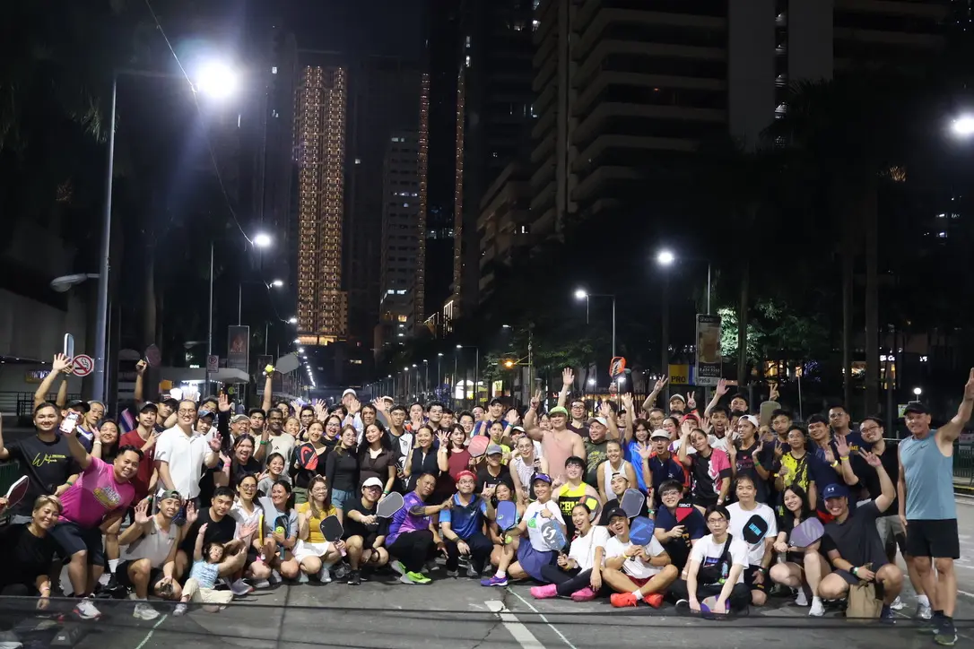 Large group of people posing together on a city street at night, with tall illuminated buildings in the background and many waving hands.