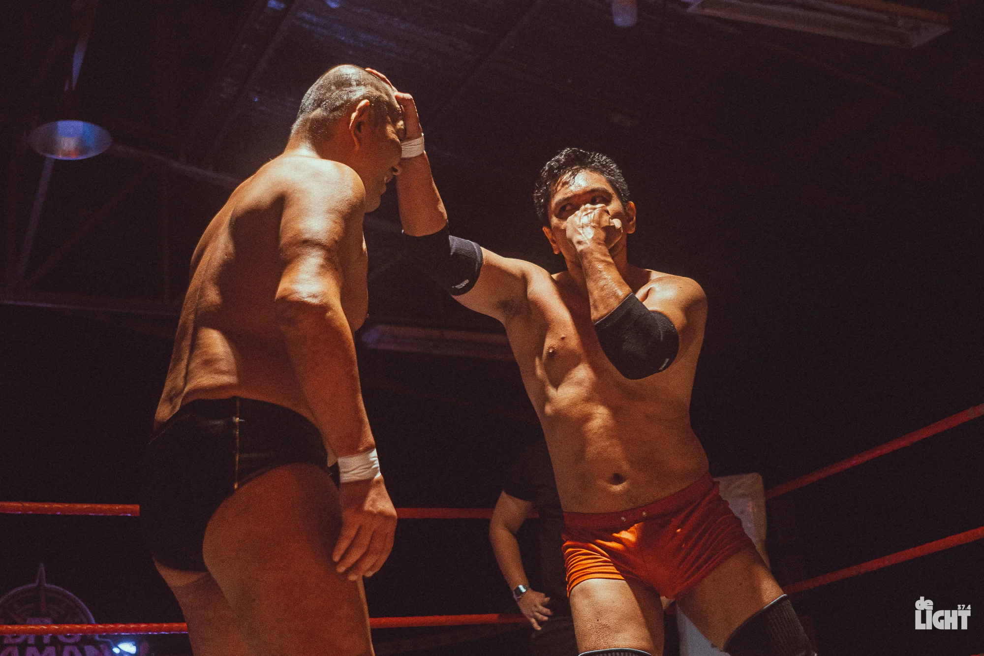 Suzuki vs ravena - the game Two shirtless wrestlers in a ring: one delivering a forearm strike to the other's head while the other covers his face with his arm; dark arena lighting and red ropes visible in the background.