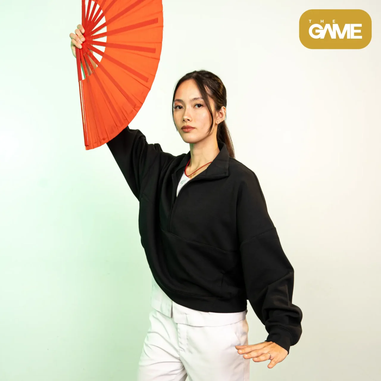 Portrait of a woman in a black jacket holding a red paper umbrella, with The Game logo in the top-right corner.