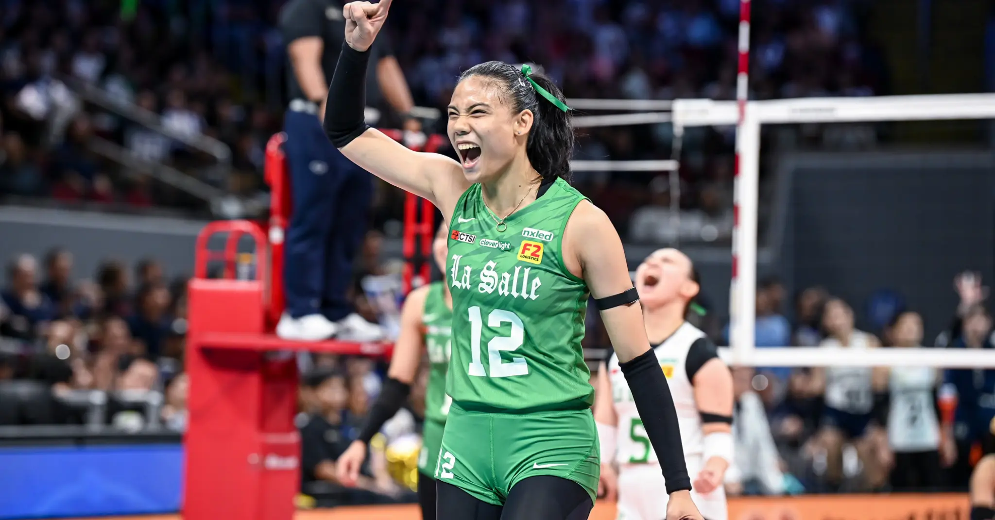 Female volleyball player in a green La Salle jersey #12 raises her fist in celebration on a packed indoor court, crowd in the stands behind her.