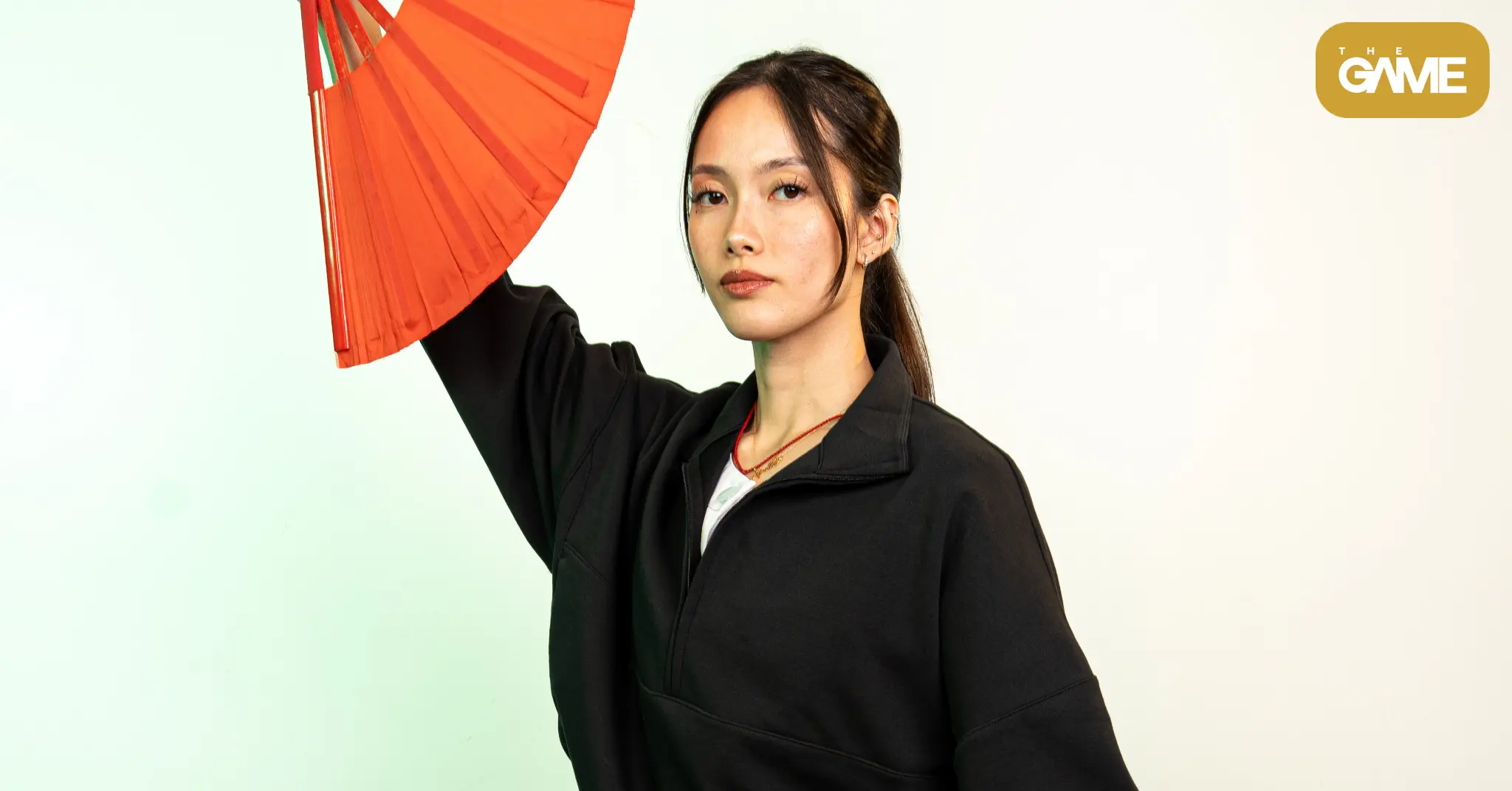 Portrait of a woman in a black jacket holding a red paper umbrella, with The Game logo in the top-right corner.