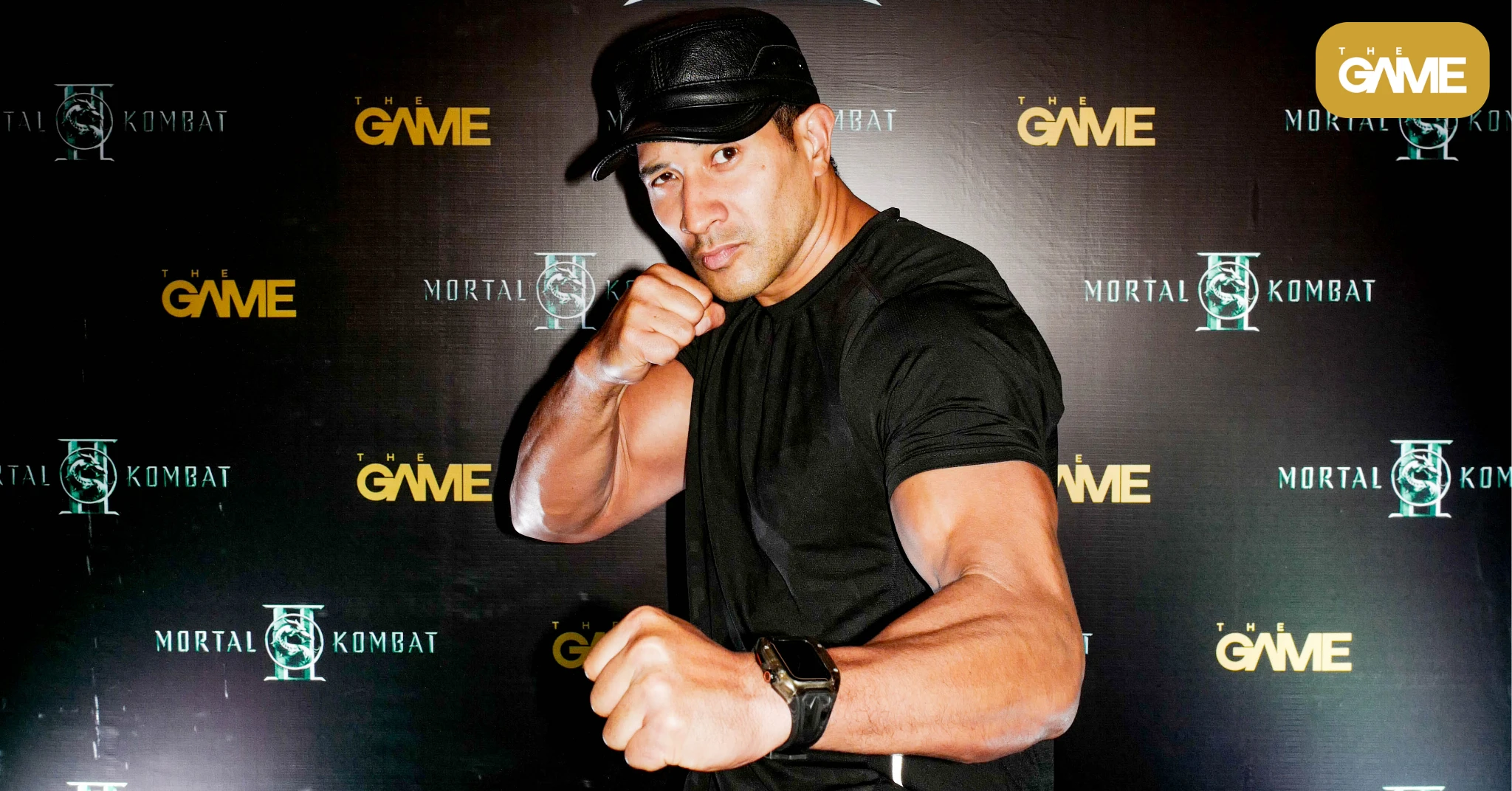 Man in a black shirt and cap poses with a clenched fist toward the camera at a Mortal Kombat/The Game backdrop.