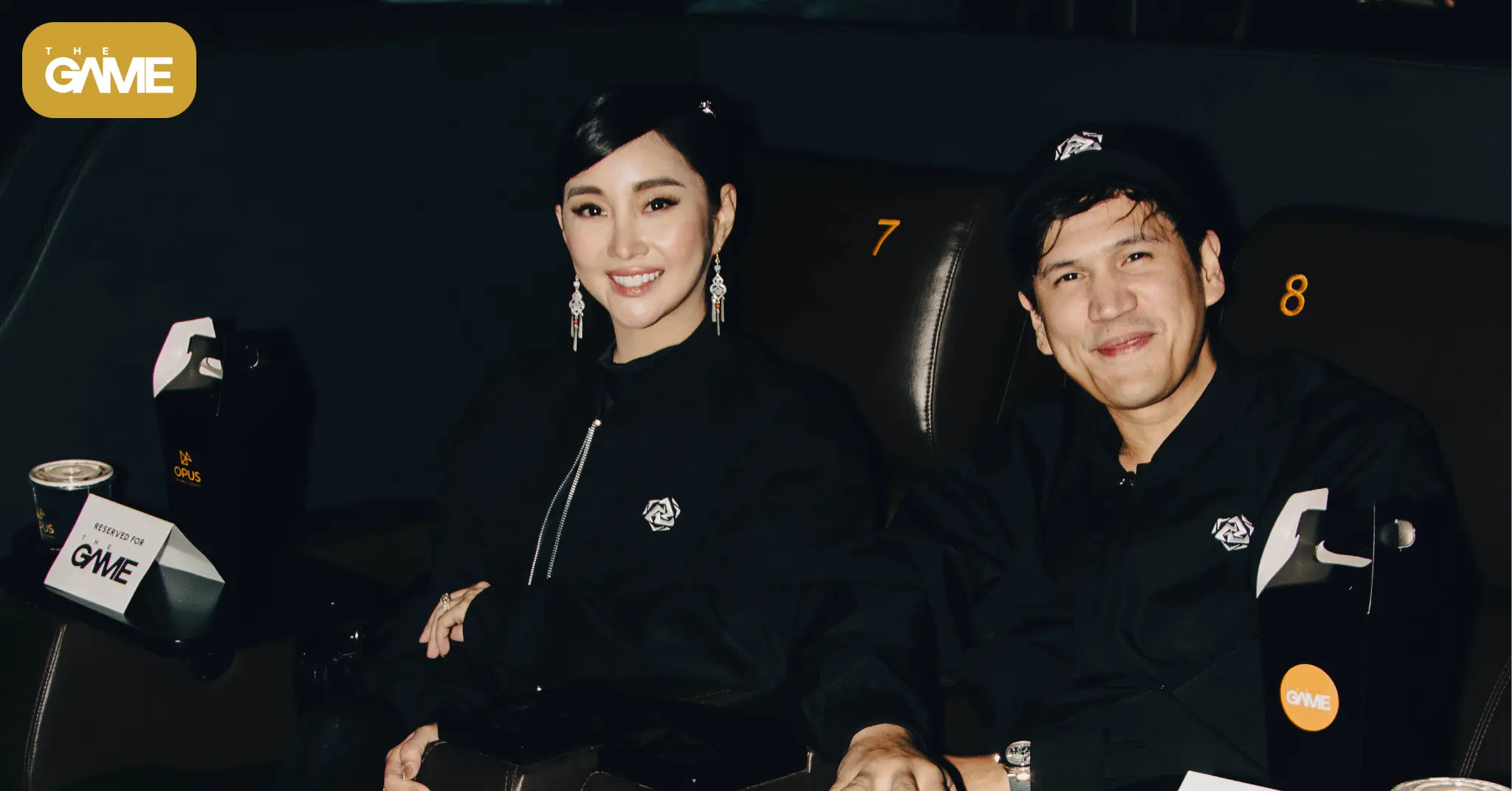 Two people sit in dark theater seats at a GME event, smiling at the camera; woman on the left with earrings, man on the right. Logo visible in the top-left corner.