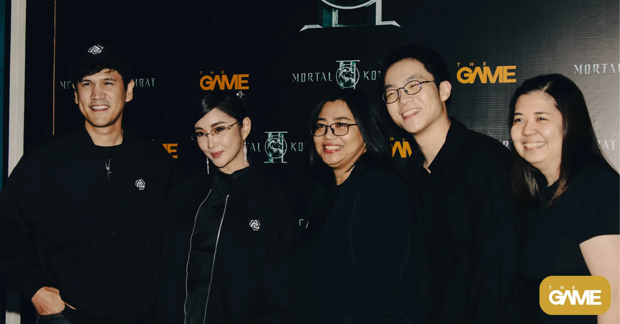 Five people in black outfits posing for a photo in front of a The Game backdrop with logos on a dark wall.
