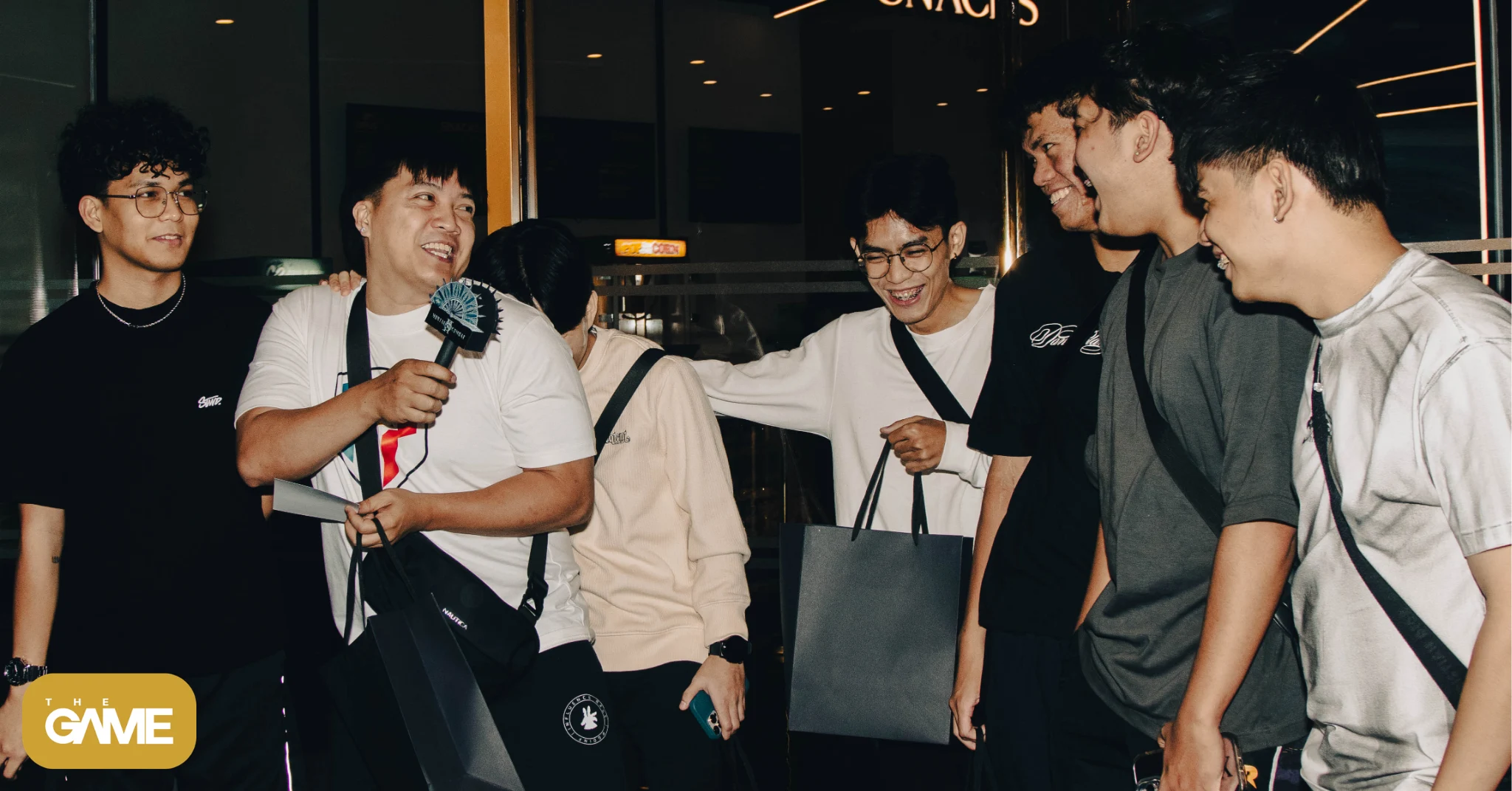 The game watermark banner wide - the game Six people standing together at a night event; one man holds a microphone and paper while others smile and pose with bags in a casual group photo.