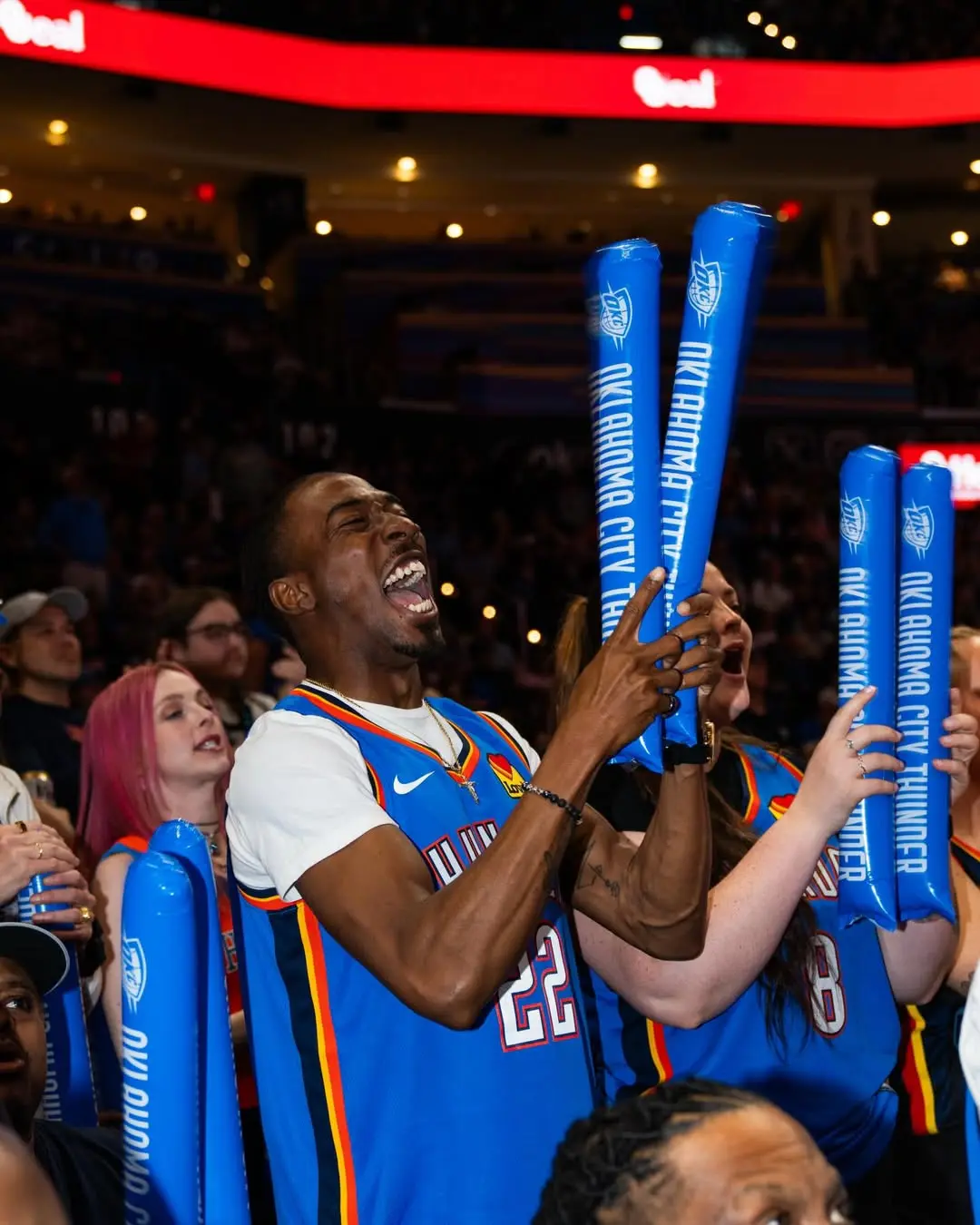 Excited basketball fans in blue Oklahoma City Thunder jerseys cheer and wave blue foam sticks in a packed arena.