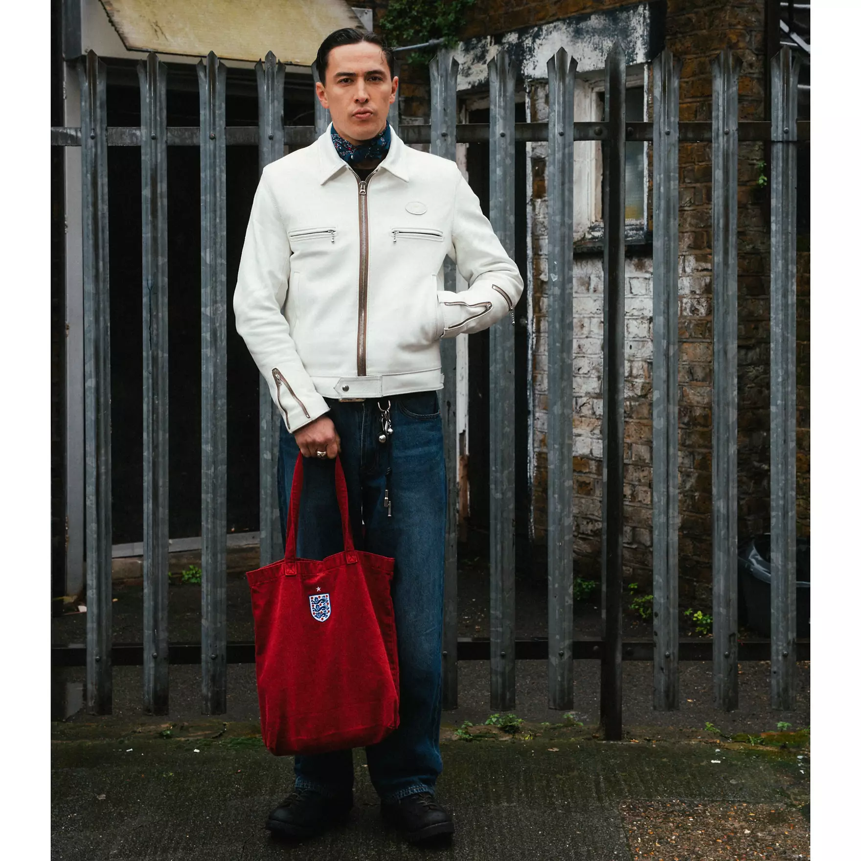 Tote - the game Man wearing a white jacket and jeans stands in front of a metal fence, holding a red tote bag with a small emblem.