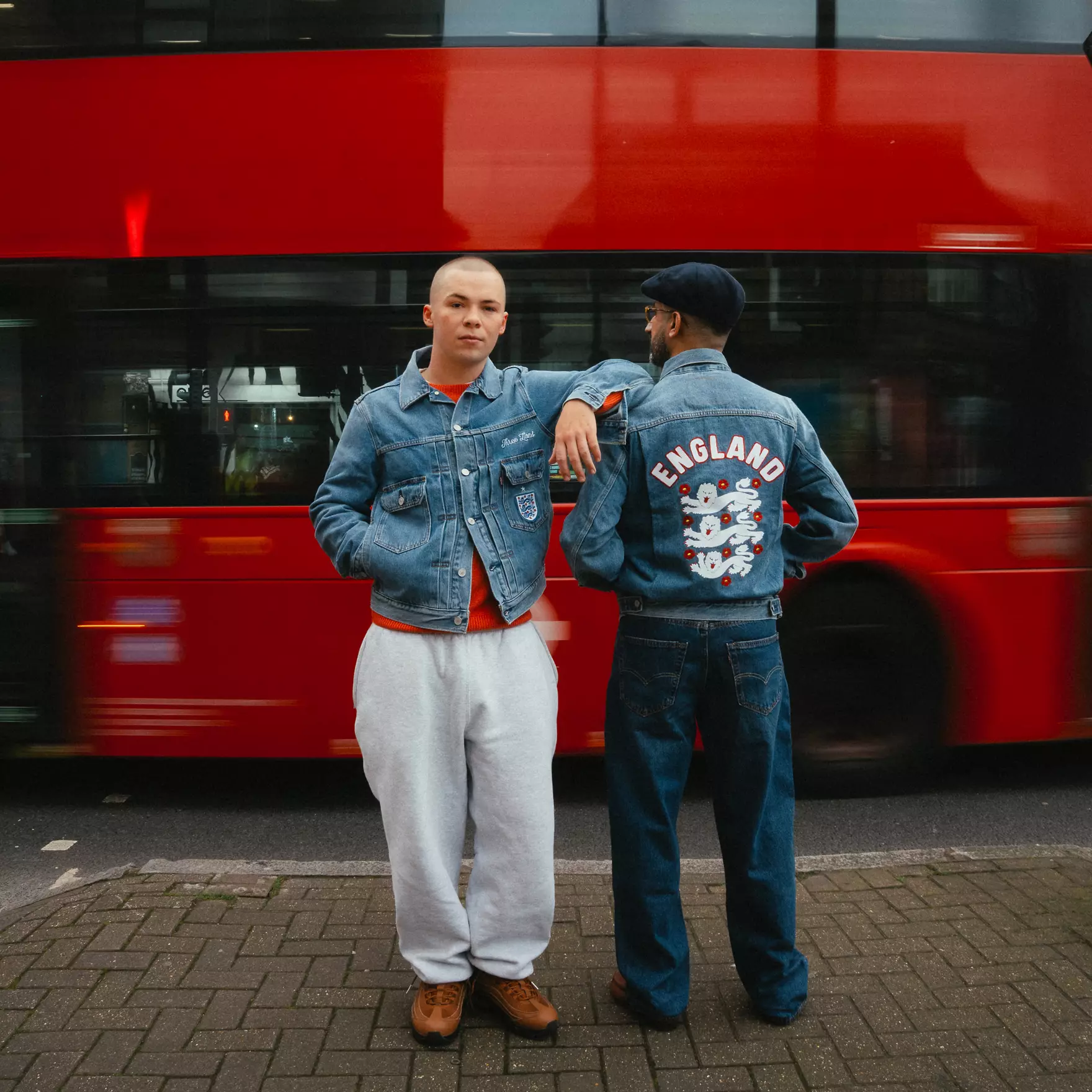 Type ii - the game Two men in denim jackets posing in front of a moving red bus on a city street; one faces the camera, the other shows an England jacket with white lions on the back.