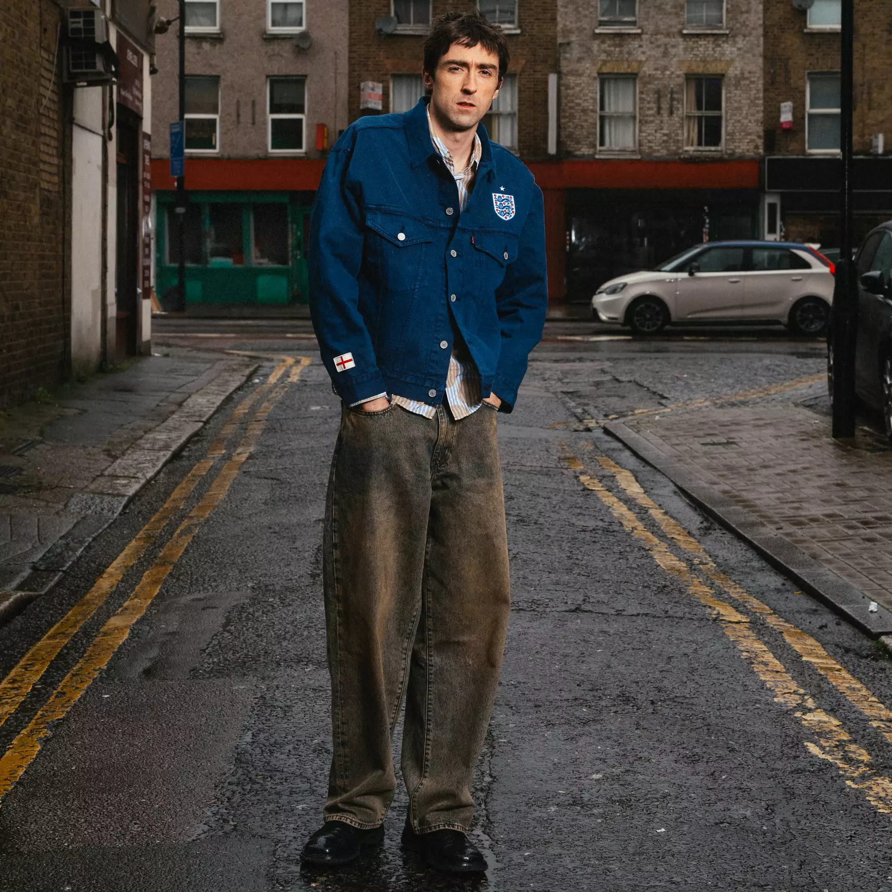 Type iii - the game Man in a blue England jacket with crest stands in the middle of a wet urban street, hands in pockets.