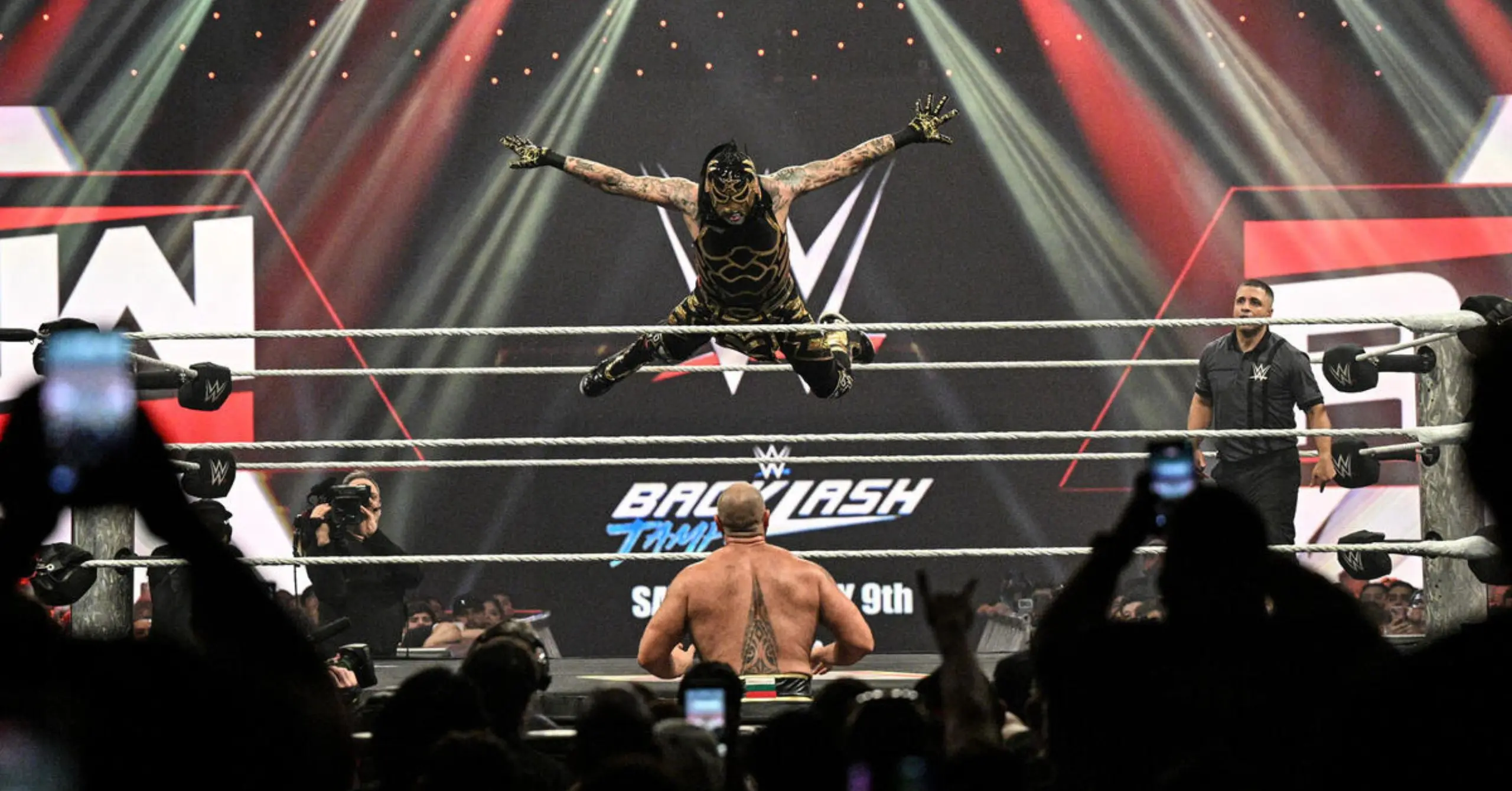 Wrestler in black and gold mask mid-air performing a jump in a WWE ring, crowd cheering around him