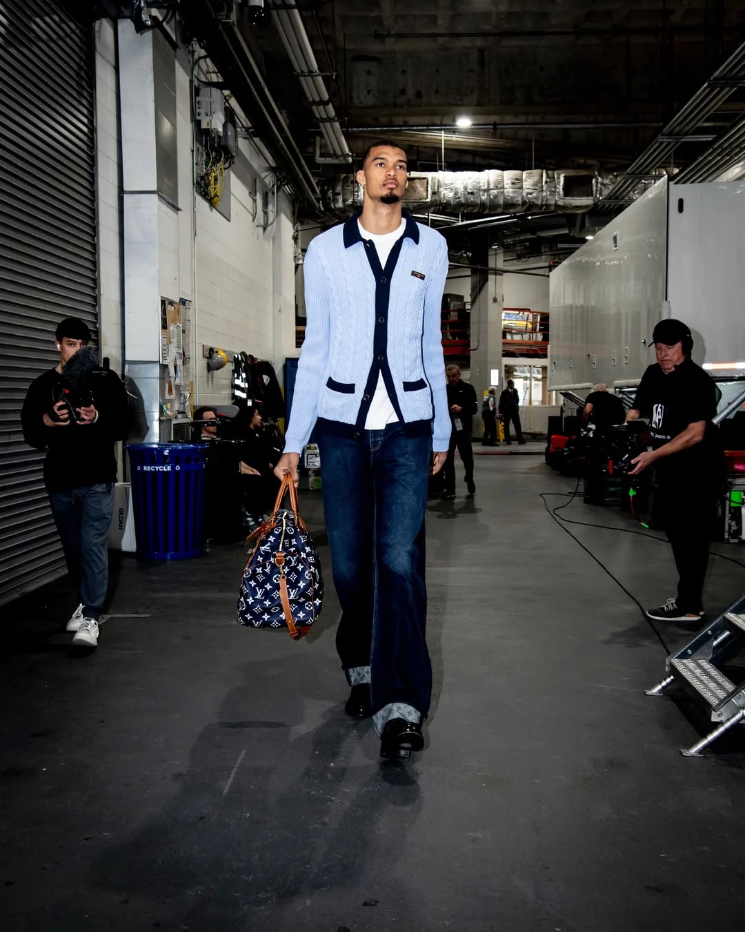 Victor wembanyama - the game Model backstage in a light blue cardigan with navy trim, white shirt, dark jeans, carrying a patterned duffel bag in a tech-filled corridor.