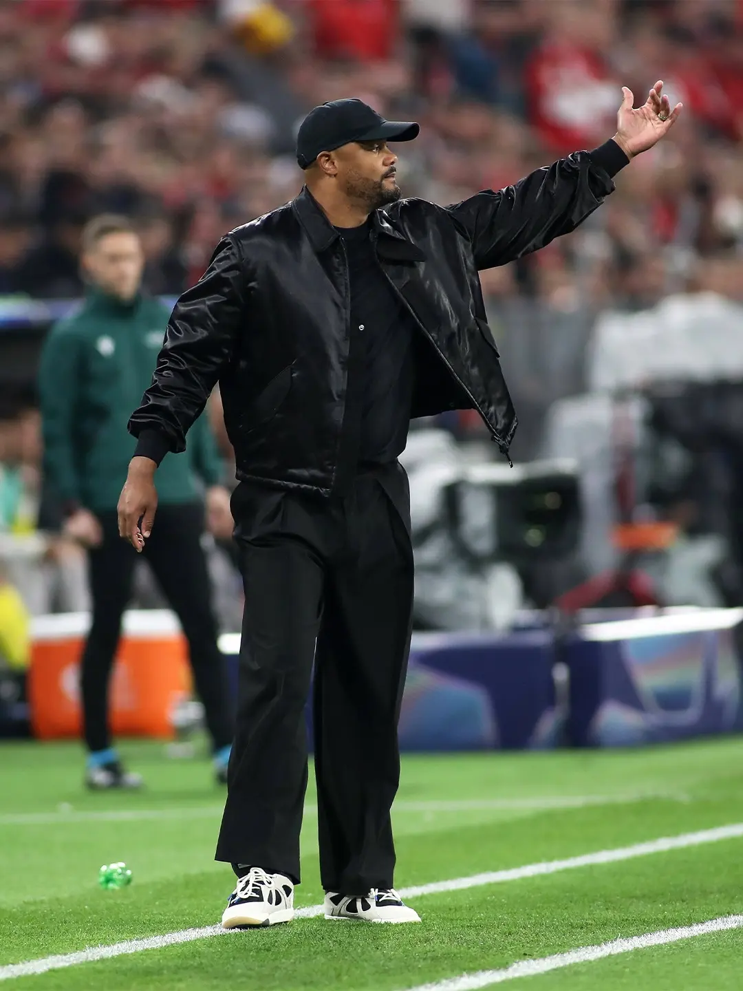 Vincent kompany - the game Man in a black jacket and cap on the sideline of a football pitch, raising his right hand as if signaling to players.