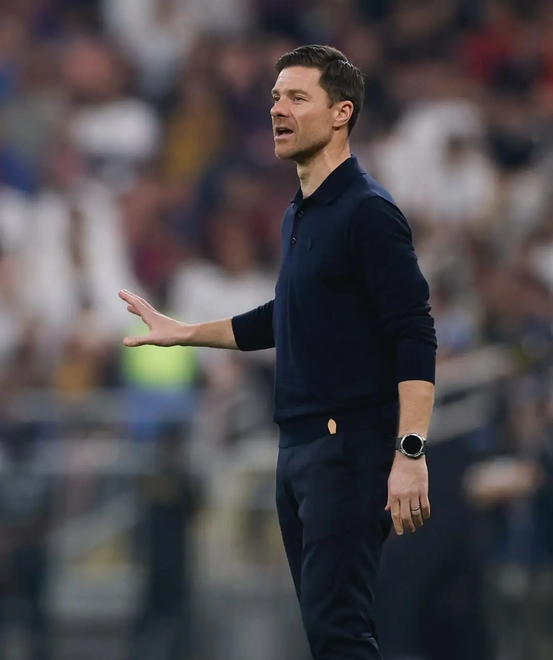 Xabi alonso - the game Coach wearing a navy polo gesturing with his left hand on a crowded stadium field behind him.