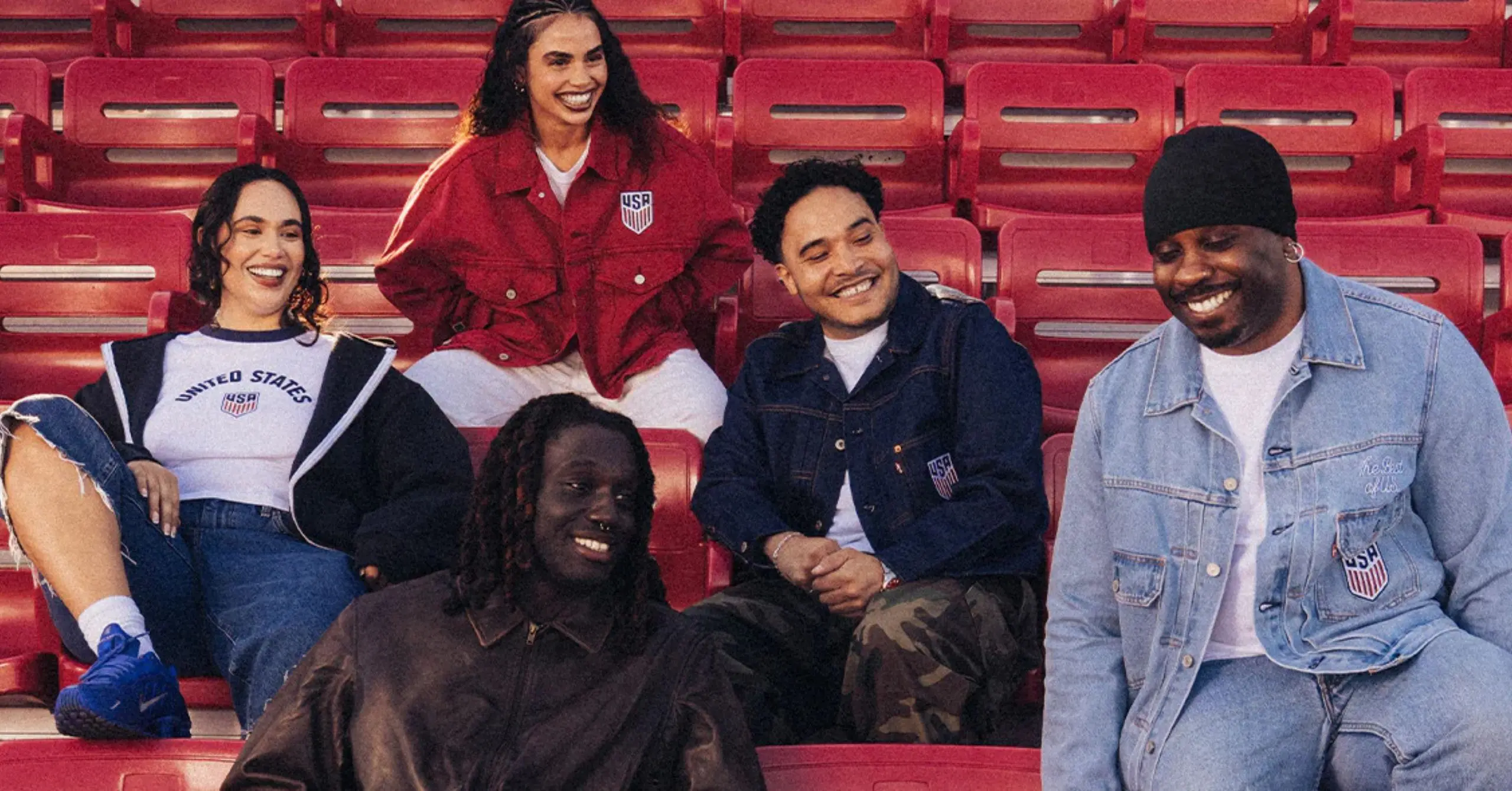 A group of friends smiling and sitting together on red stadium seats, wearing USA jackets and casual clothing.