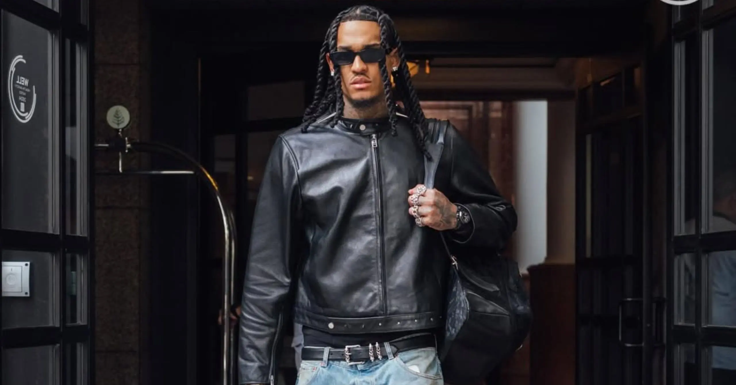 Man wearing a black leather jacket and sunglasses steps out of a building, hands on a strap of a bag, confident stride.